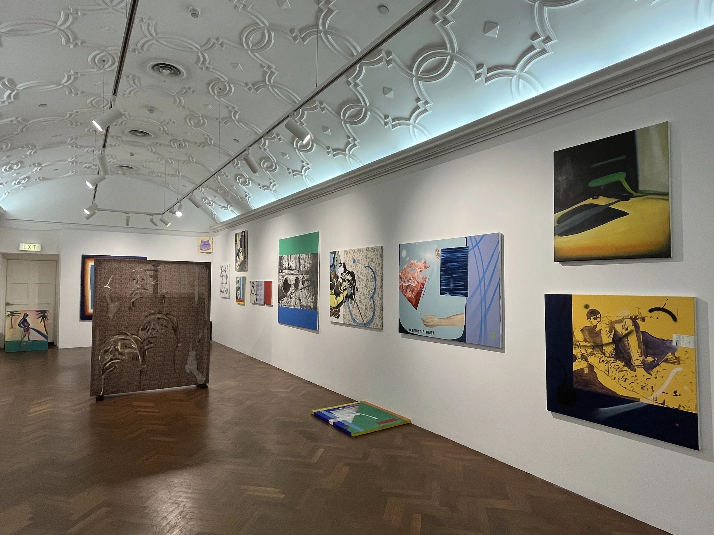Art gallery with various paintings on the wall and a large ornate ceiling, wooden floor, and low lighting.