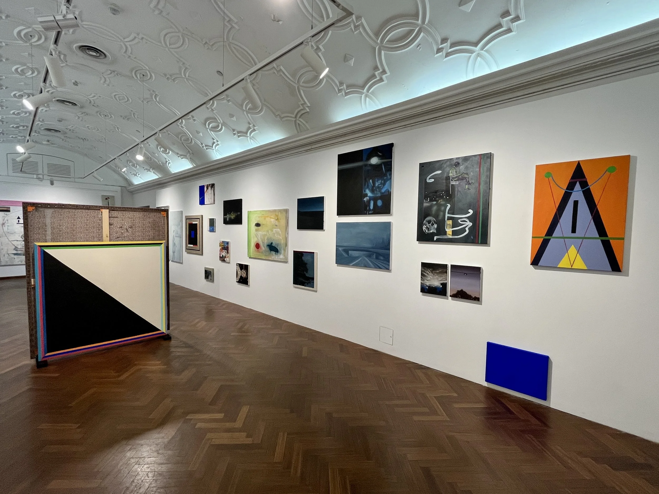 An art gallery featuring various abstract paintings on white walls and a carpeted floor.