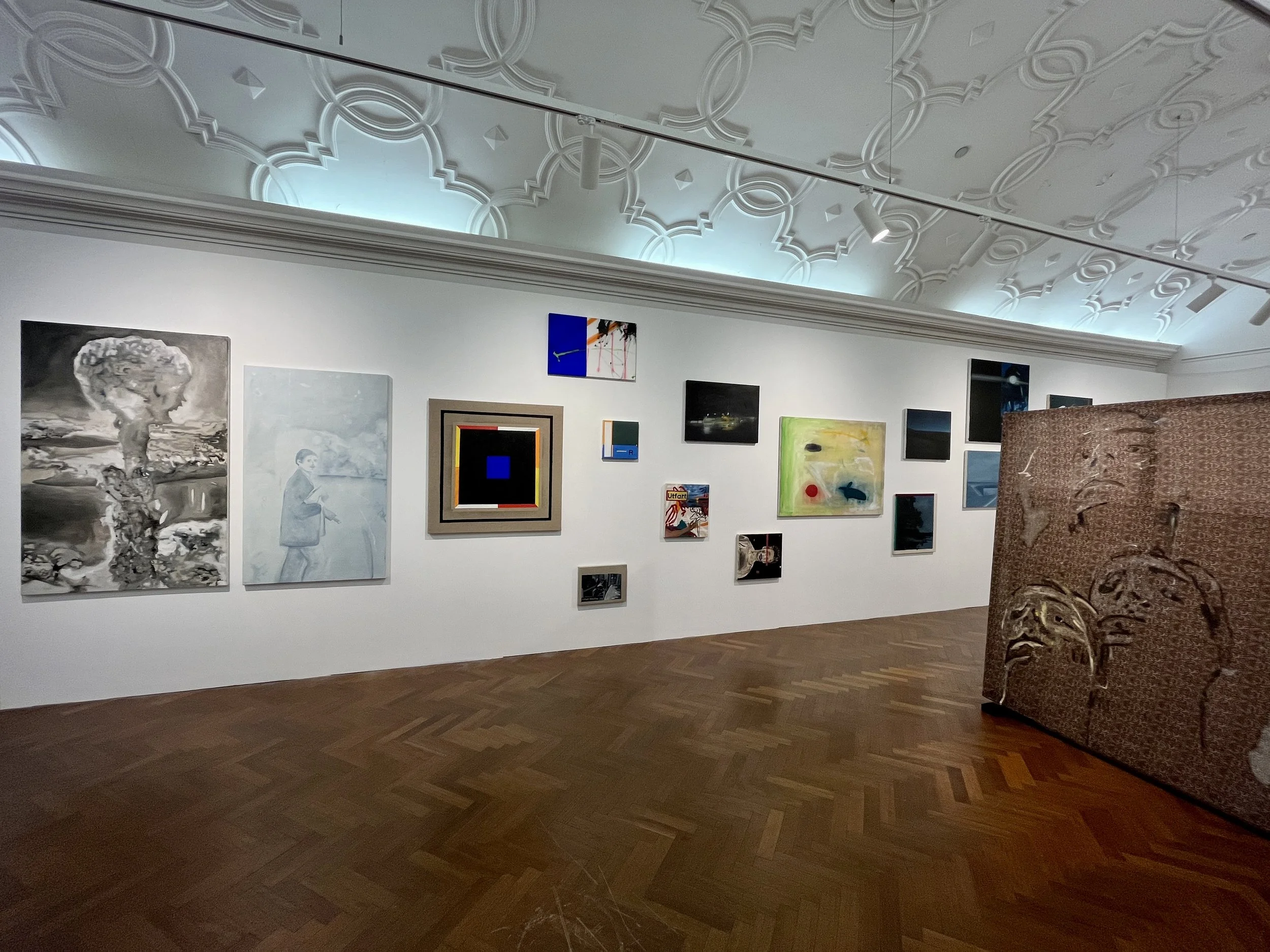 Multiple paintings displayed on a white wall in an art gallery, with a decorative ceiling and wooden floor, and a patterned chair partially visible in the foreground.