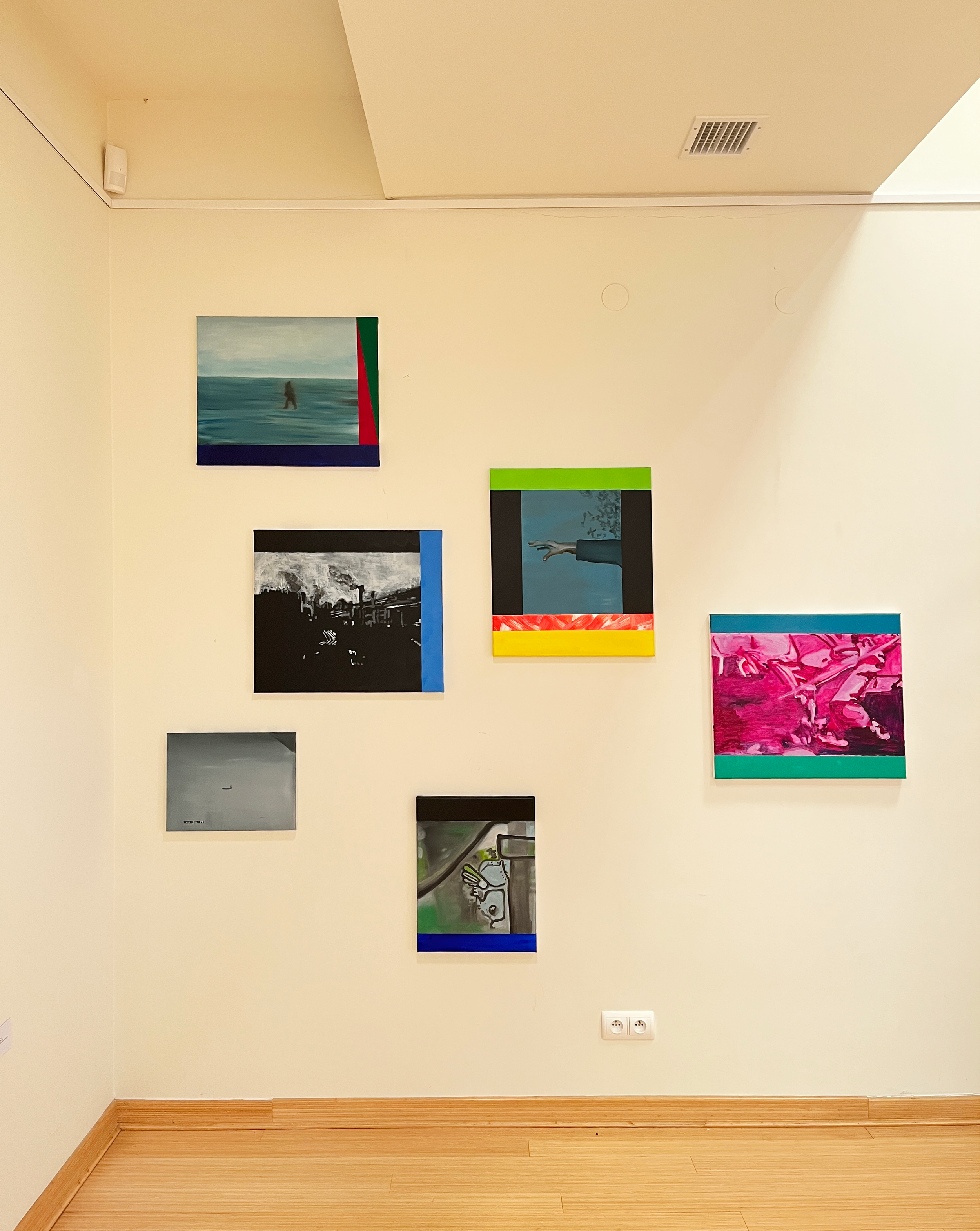 A gallery wall with various small paintings, including an ocean scene and a surreal image of a dog with a rainbow background, on a cream-colored wall with wooden flooring and two electrical outlets.