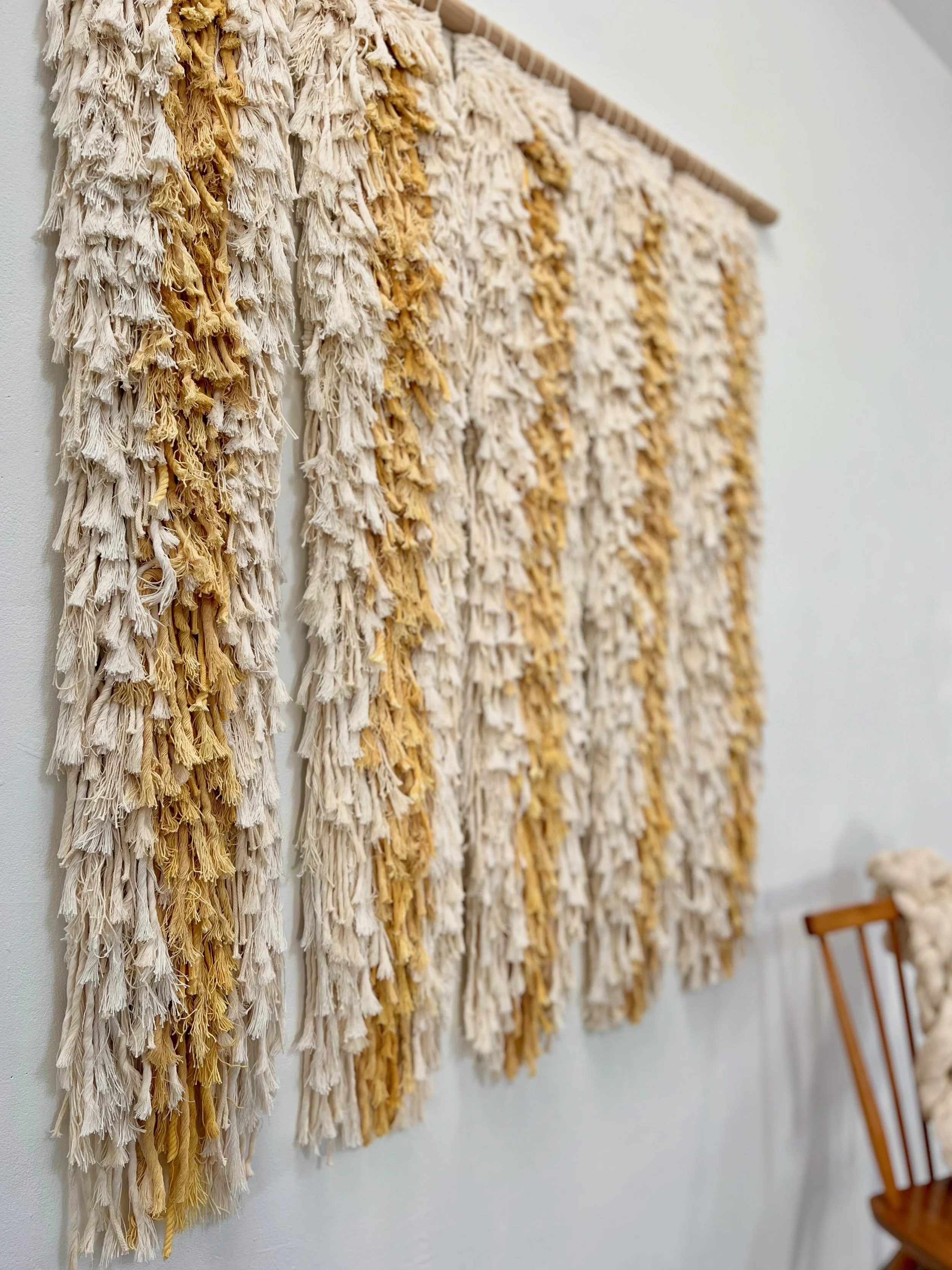 Close-up of a textured wall hanging made of cream and yellow yarn on a wooden dowel, hanging on a white wall, with a wooden chair partially visible to the right.