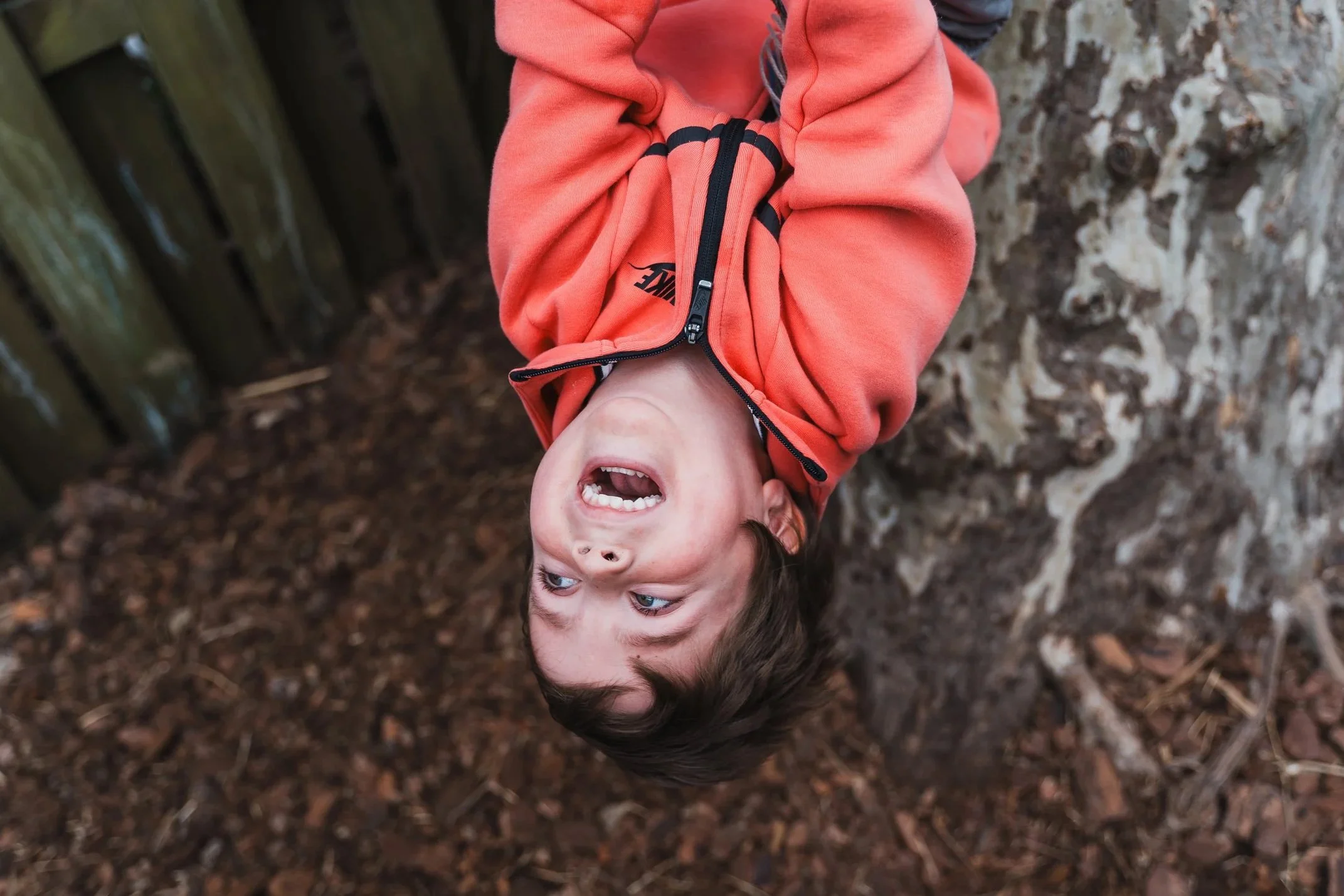 A young child with curly hair and light-colored eyes, yelling or crying outdoors with a background of trees and leaves.