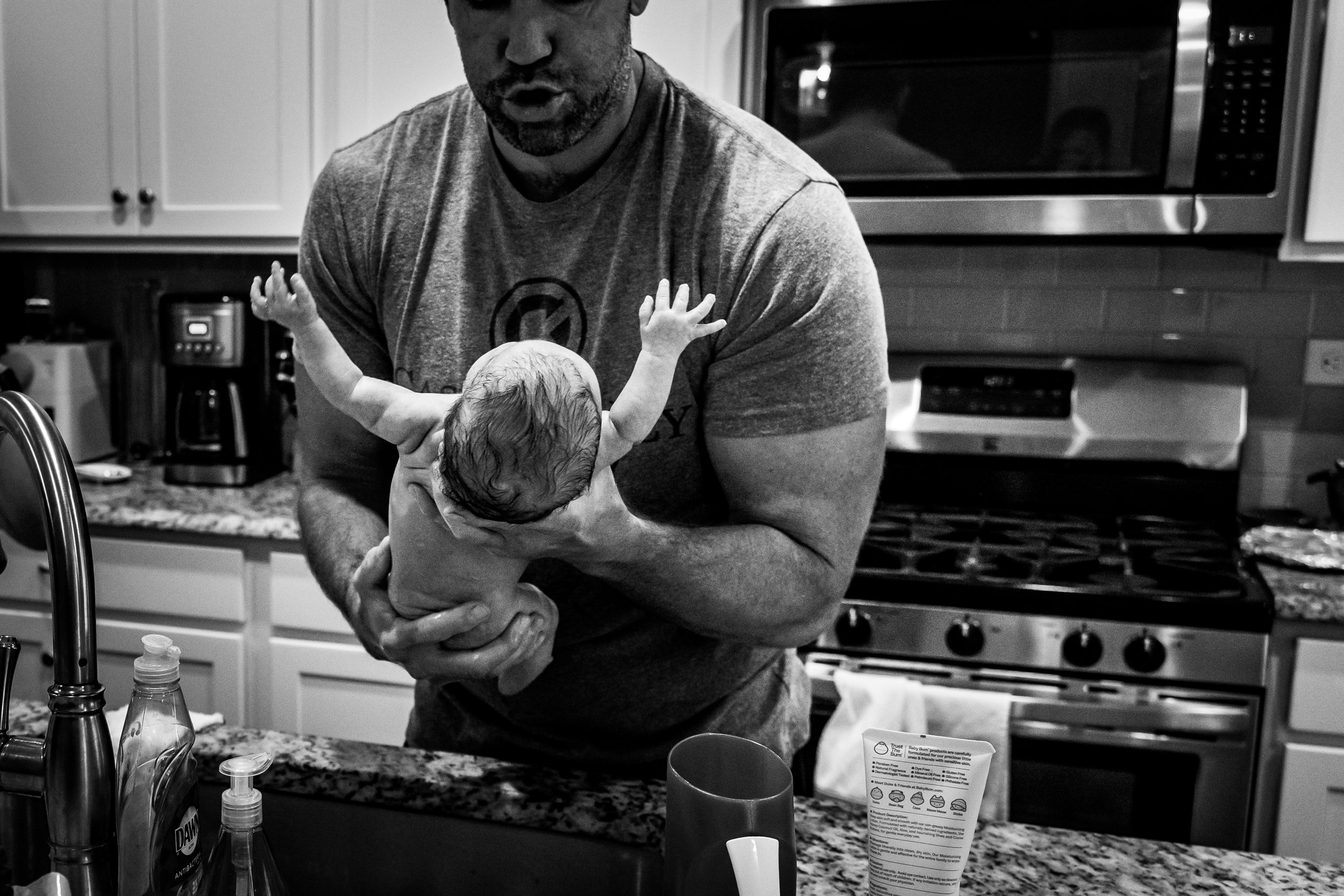 Baby getting first bath in the kitchen sink, crying, and dad is holding her, shushing