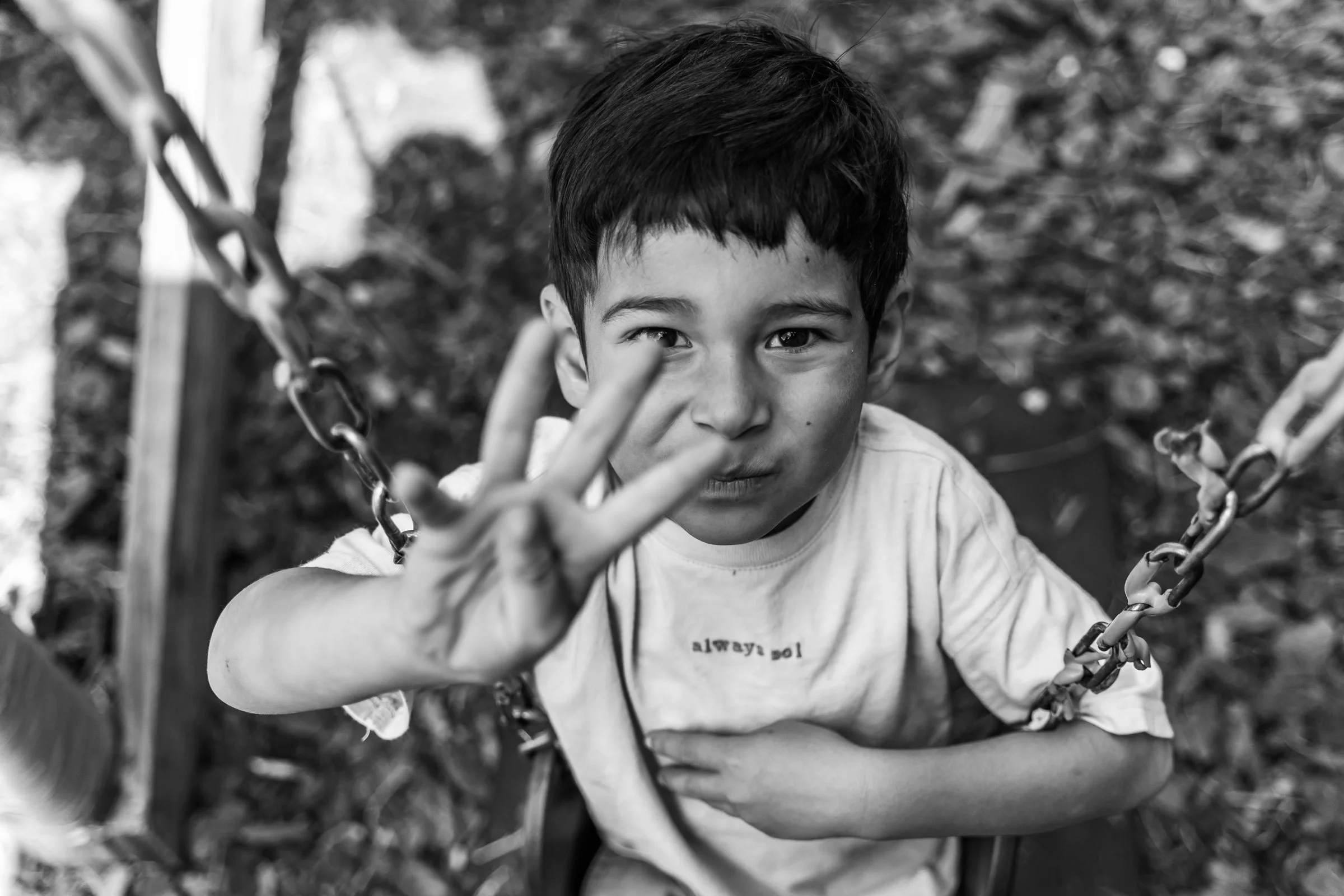 A young girl with curly hair wearing a floral shirt making a peace sign with her fingers in front of the camera, outdoors with a blurred background.