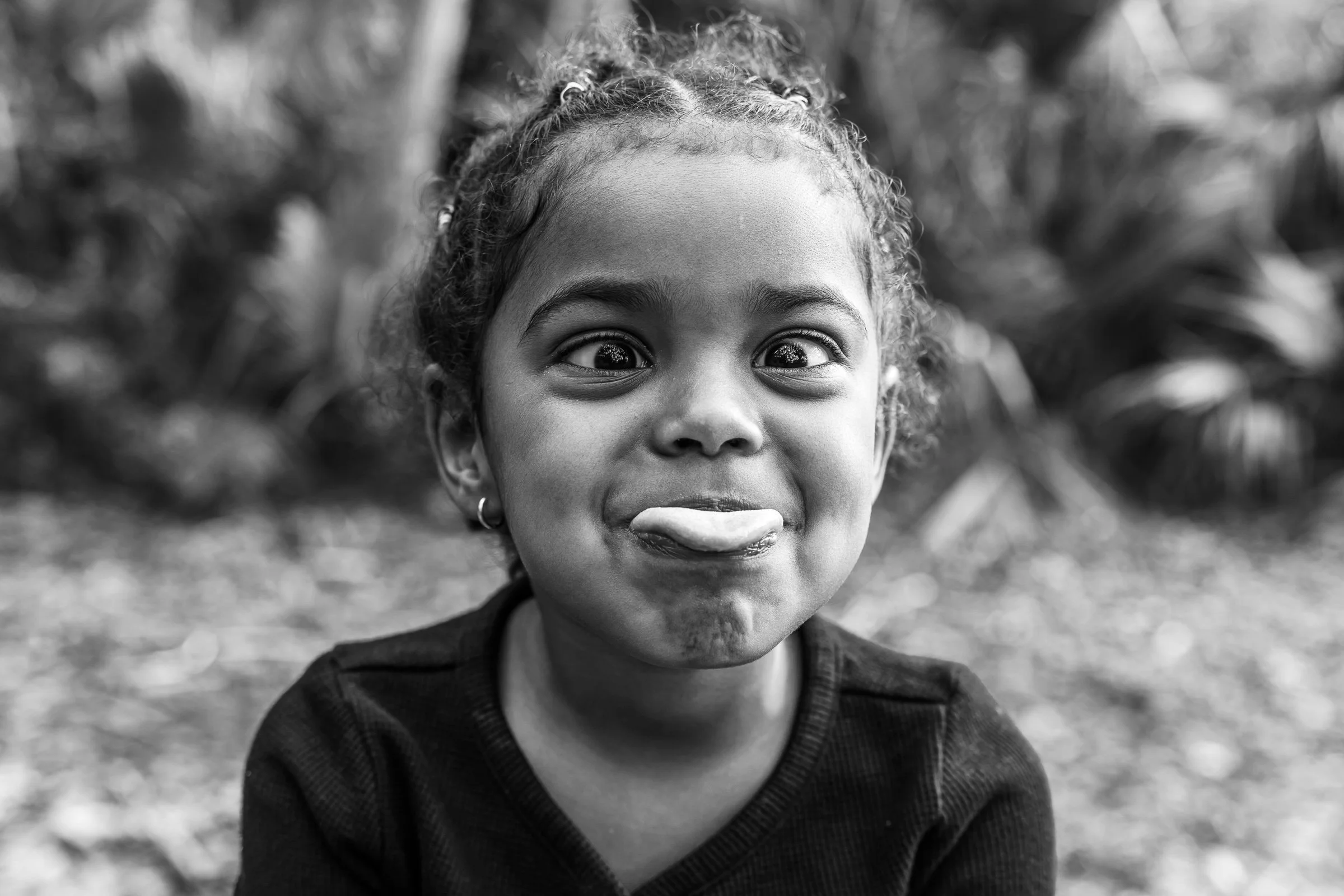 A young boy with short light brown hair and brown eyes, wearing a white T-shirt with faint green spots, standing outdoors in front of a wooden fence.