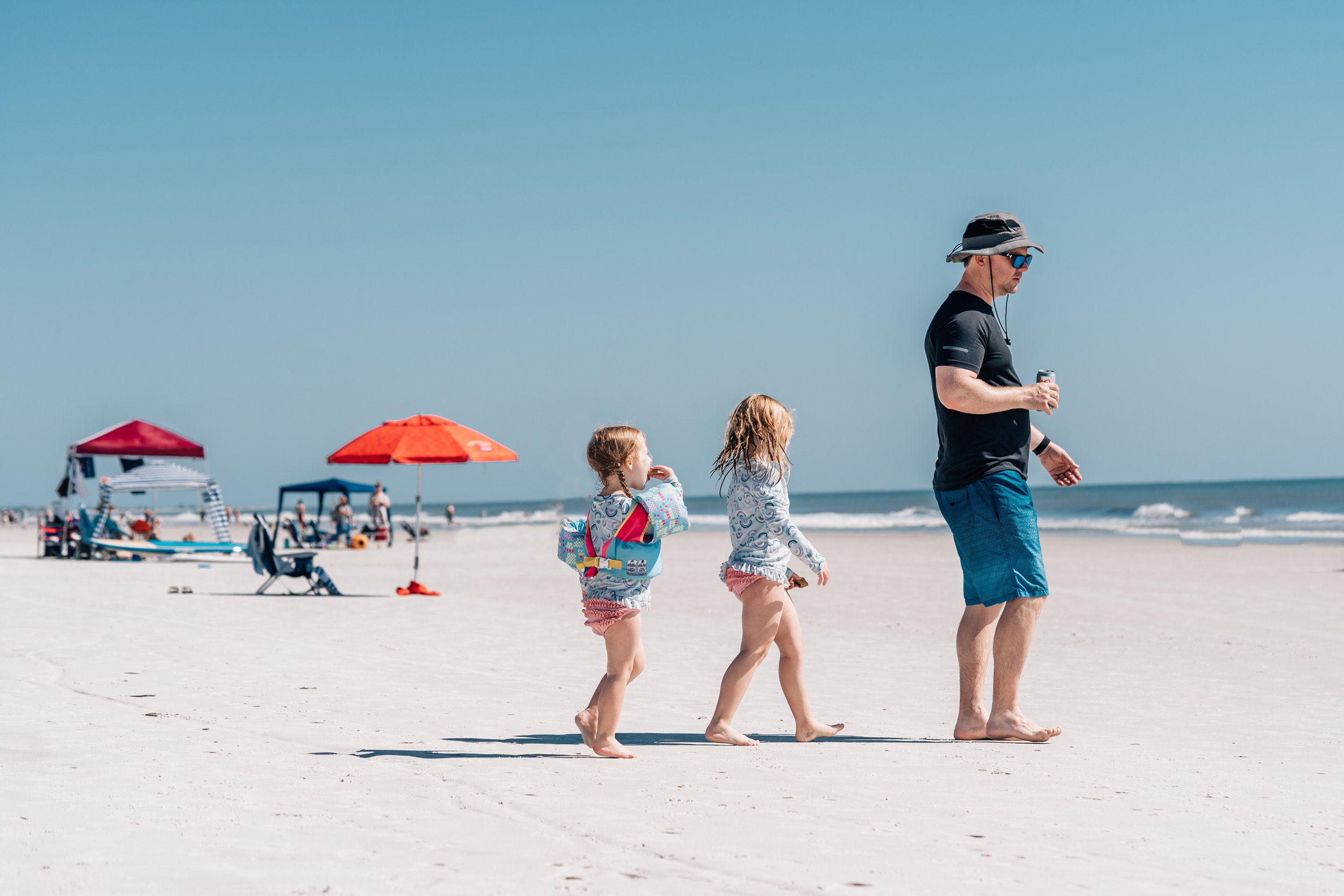 Documentary photo of two little girls following their dad, who is drinking a beer at the beach