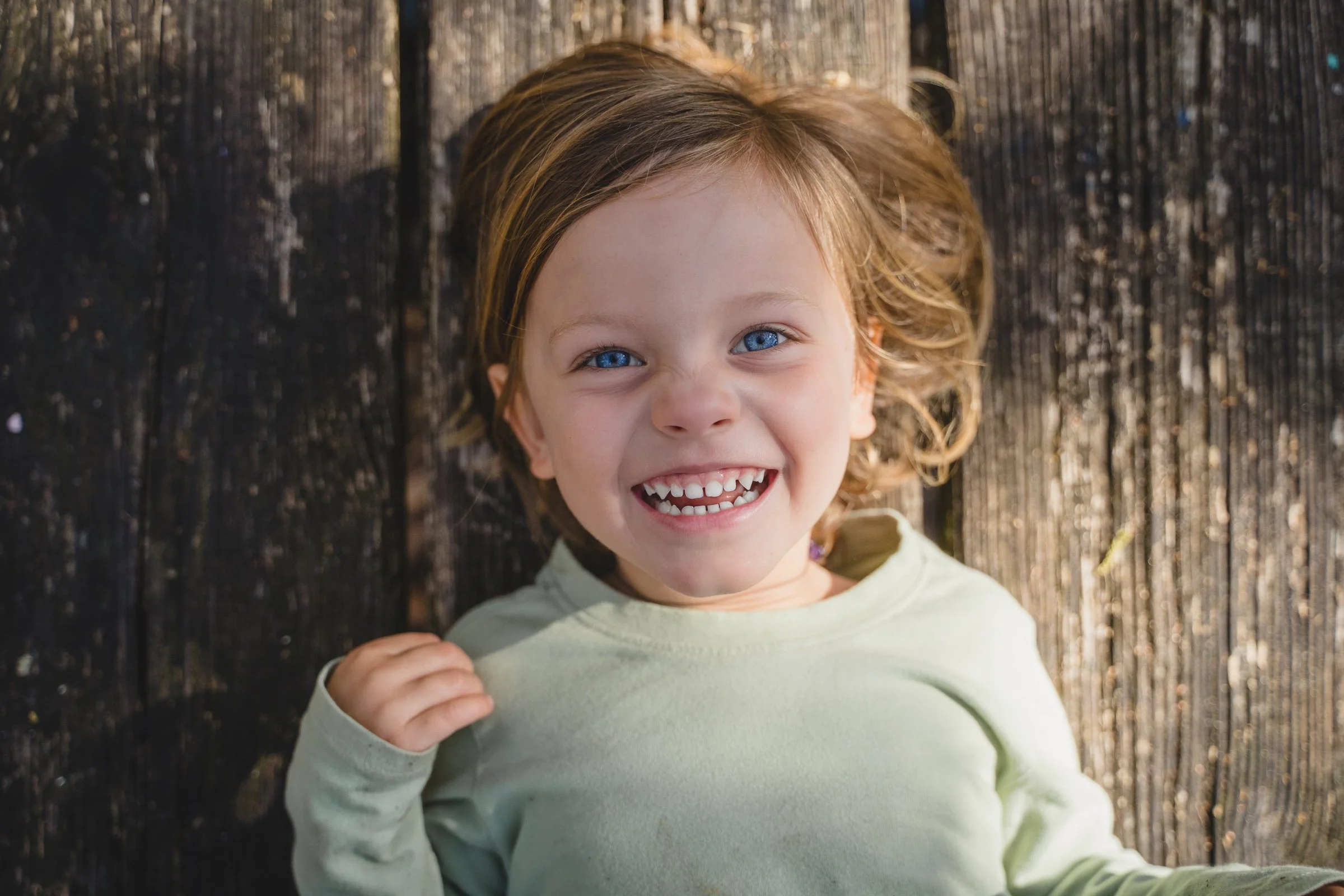 A young girl with curly blonde hair and blue eyes making a duck face while looking up at the camera, wearing a pink shirt, outdoors on a dirt surface.