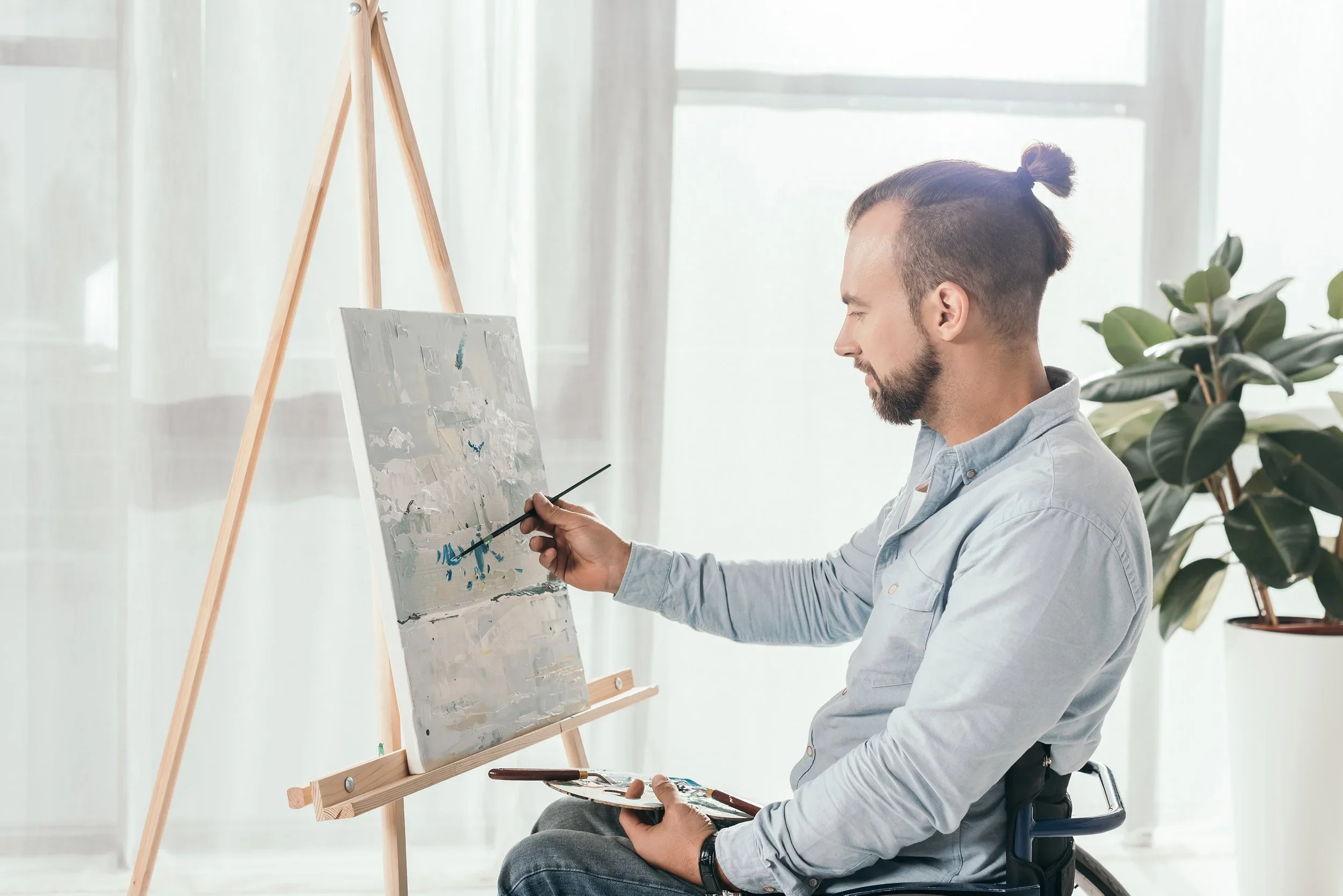 A man sitting on a chair painting on a canvas placed on an easel in a bright room with large windows and a potted plant in the background.