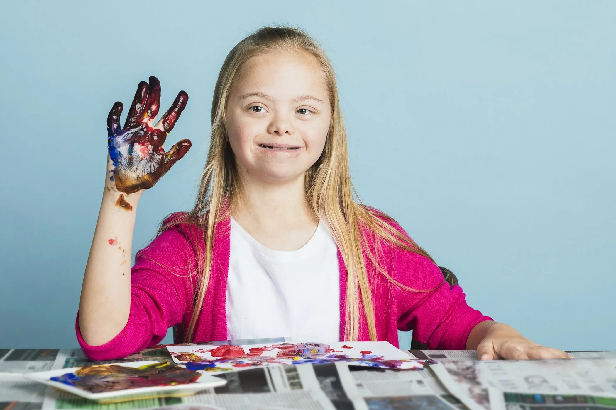 A young girl with long blonde hair, wearing a pink jacket and white shirt, is sitting at a table covered with newspapers and painting. Her right hand, covered in red, blue, yellow, and black paint, is raised and she is smiling slightly against a plai