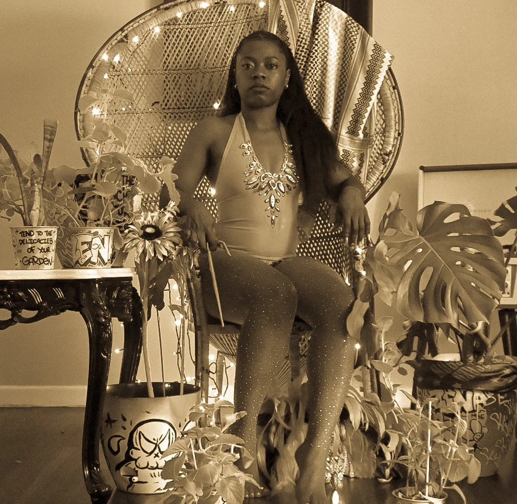 Black woman seated in a wicker chair wearing a Caribbean-inspired carnival outfit, holding a blade, surrounded by lush plants and graffiti-painted pots, symbolizing identity, power, and cultural duality.