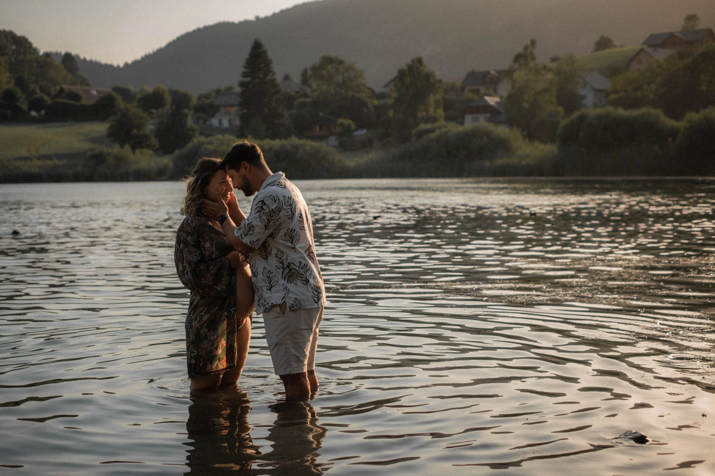 Un couple se tient dans une rivière lors du coucher du soleil, leur visage près l'un de l'autre, avec des montagnes et des maisons en arrière-plan.