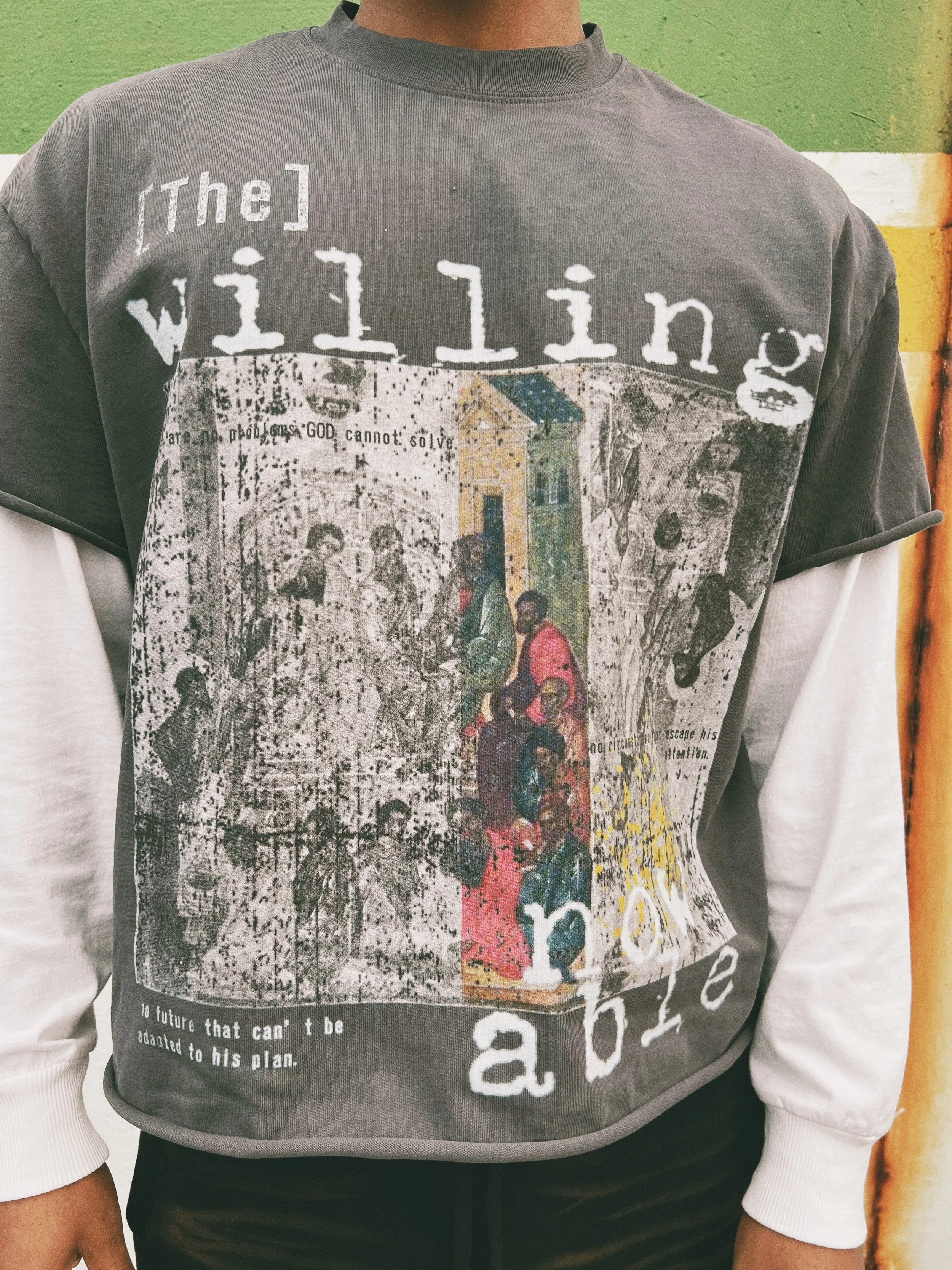 Person wearing a gray T-shirt with layered sleeves. The shirt has artistic and text elements, including the words "The Willing Now Able" and various vintage-style images. 