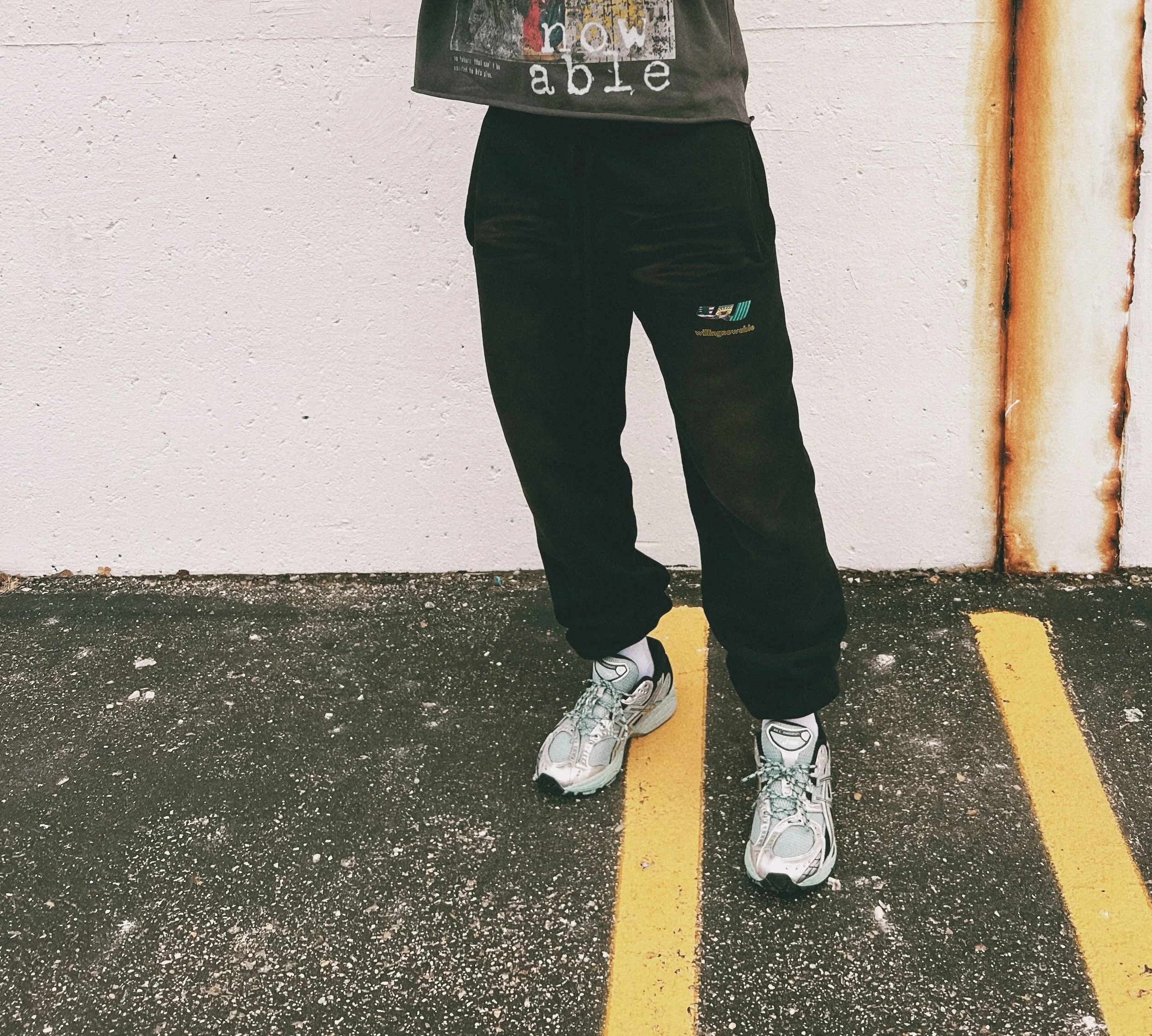 Person wearing black sweatpants, graphic T-shirt, and silver sneakers standing on pavement with yellow lines.