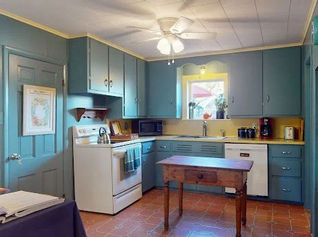 Kitchen with blue cabinets, white stove, microwave, breadbox, yellow walls, small window, ceiling fan, wooden table, and tiled floor.