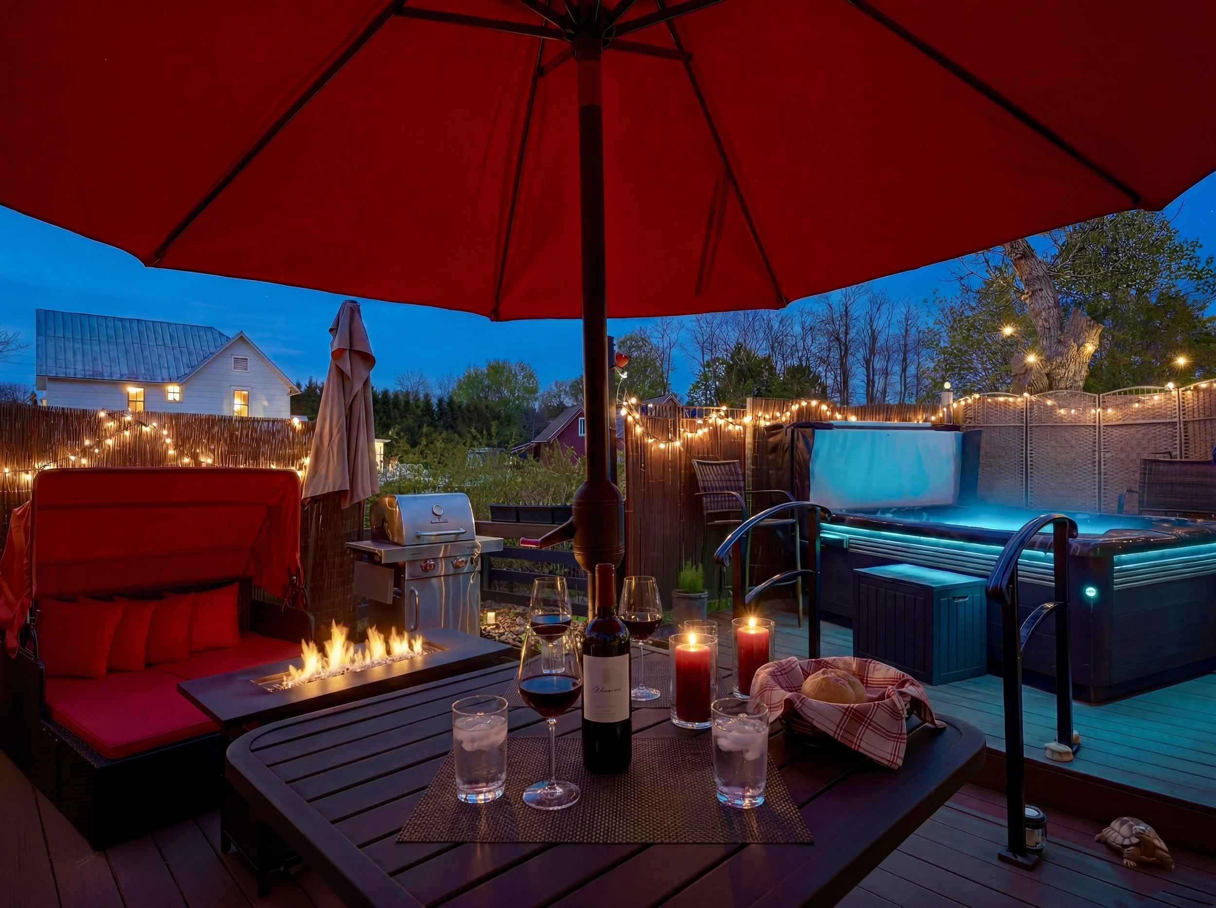Nighttime backyard patio with red umbrella, string lights, a seating area with a firepit, wine glasses, a pizza, and a hot tub, with trees and neighboring houses in the background.