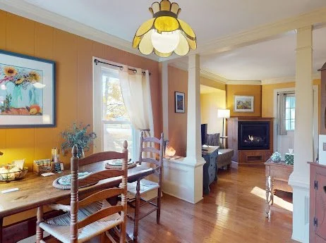 Dining room with wooden table, chairs, floral centerpiece, window with white curtains, and a living room with a fireplace and seating area.