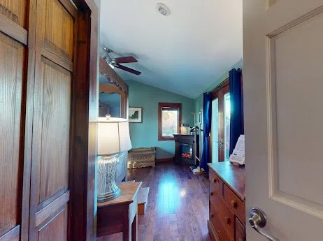 View of a cozy living room with hardwood floors, teal walls, a ceiling fan, a window with blue curtains, and furniture including a sideboard and a fireplace.