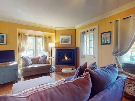 Living room with yellow walls, white curtains, a gray couch, a purple armchair, a fireplace, and a TV