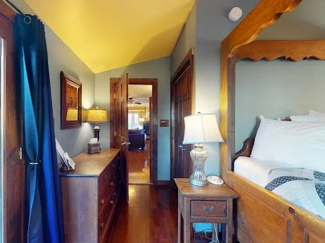 Bedroom with a wooden four-poster bed, side table with a lamp, blue curtains, and a dresser facing the hallway.