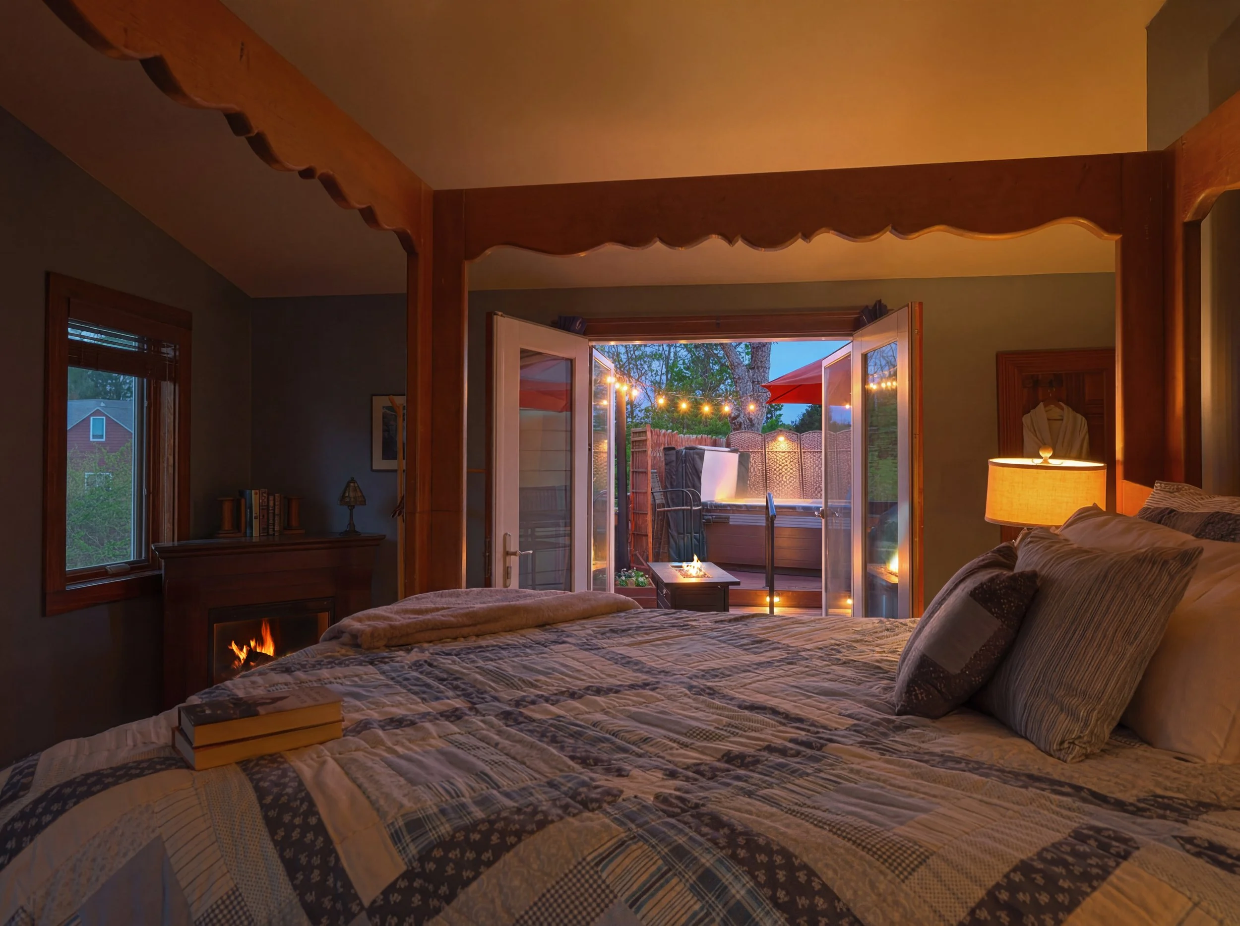 A bedroom with a bed covered in a patchwork quilt, a window with blinds, a fireplace, a wooden bookshelf, a desk lamp, and open double glass doors leading to an outdoor patio with string lights and a red umbrella.