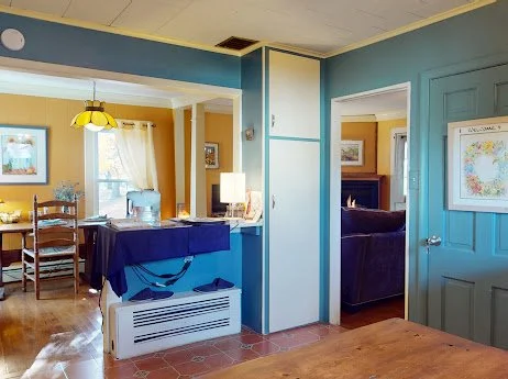 Living room with blue walls, a dining area with a wooden table, a kitchen with a blue counter, and a hallway with a door leading to a bedroom with a dark-colored bed.