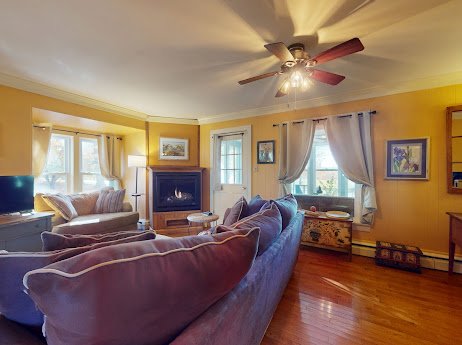 Living room with yellow walls, a ceiling fan, a fireplace, and multiple windows with curtains, furnished with sofas and a TV.