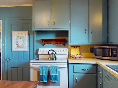 Kitchen with blue cabinets, a white stove with a kettle, a microwave, and teal towels hanging on the oven handle.