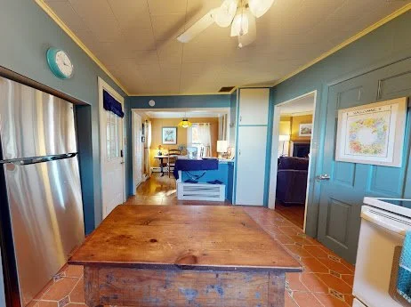 Interior view of a kitchen leading to a dining area with a view into a living room, featuring wooden floors, blue walls, and a ceiling fan.