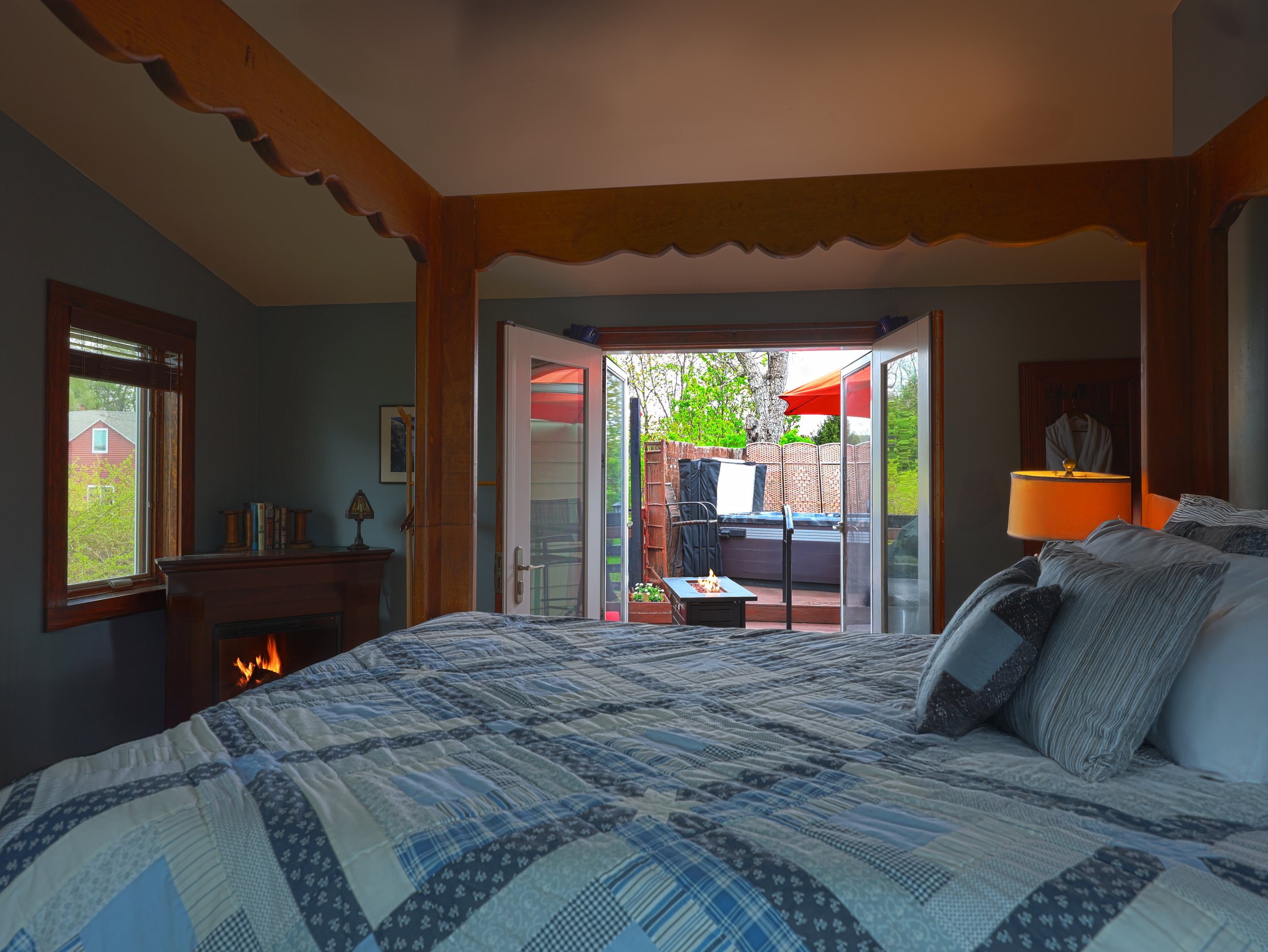View from inside a bedroom showing a bed with a blue quilt, windows, and an open door leading to a backyard patio with outdoor furniture and a red umbrella.