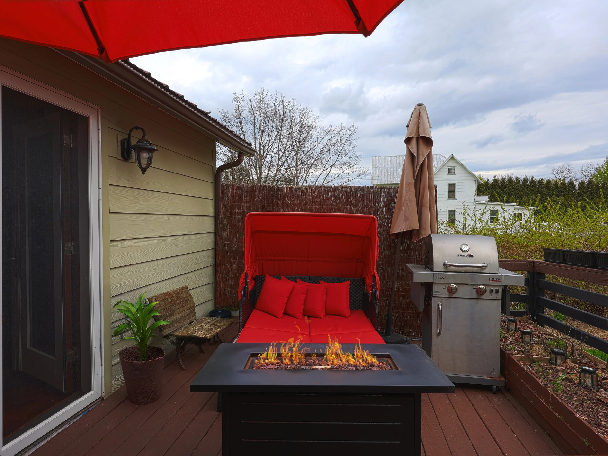 A backyard patio with a fire table, a red swing chair with red cushions, an umbrella, a grill, a potted plant, and a wooden fence, with overcast sky and neighboring houses in the background.