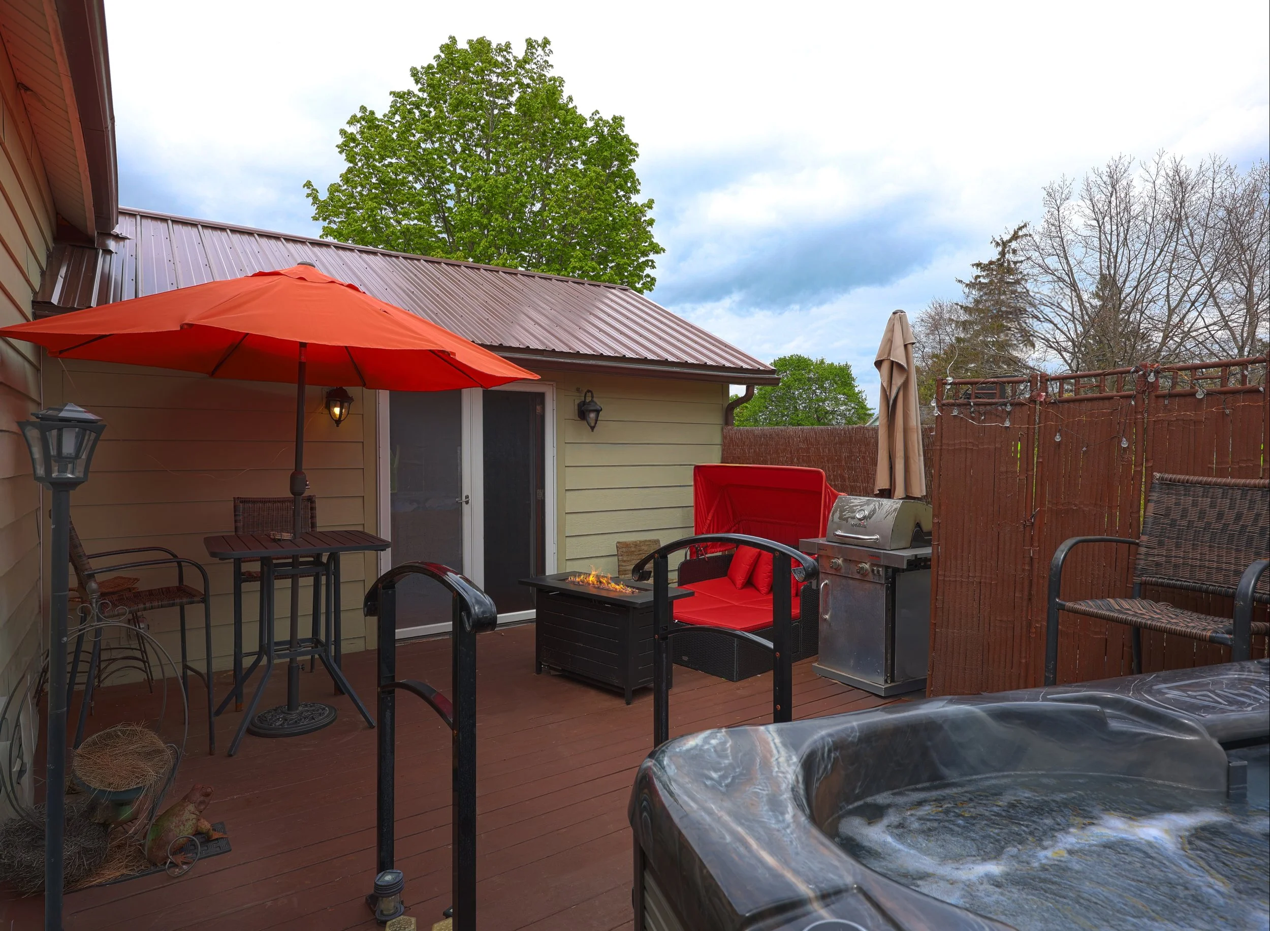 A backyard patio with a red umbrella, outdoor furniture including chairs and a sofa, a barbecue grill, string lights, and a fenced area with trees and a cloudy sky.