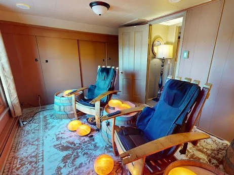 Two wooden rocking chairs with blue cushions are in a cozy room, surrounded by orange decorative lights on the floor. A plaid ottoman and a floor lamp are in the background.
