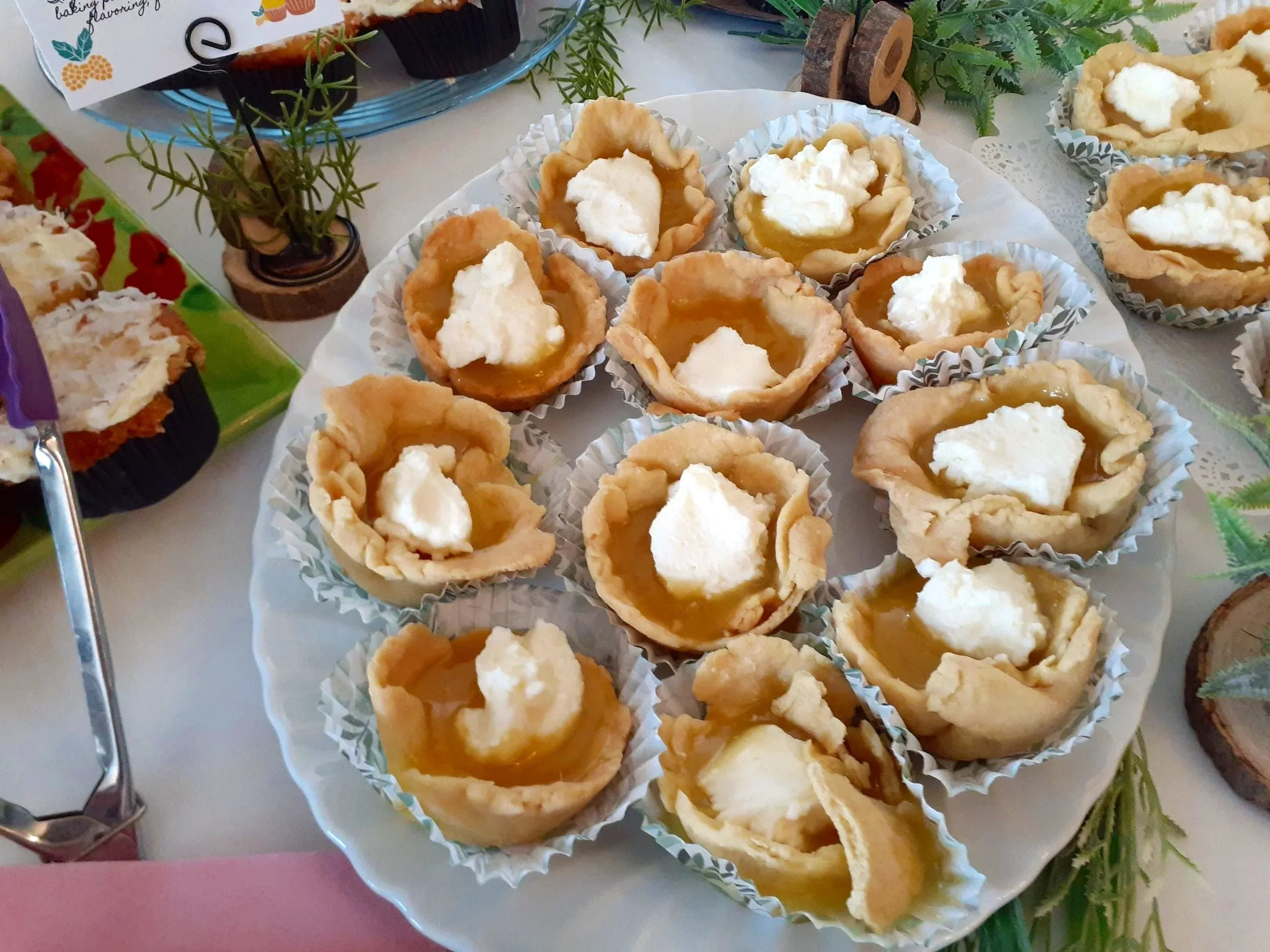 A white plate with small tartlets filled with a  home made lemon curd and dollops of whipped cream on top. The tartlets have crimped edges and are placed in paper cups. The plate is on a table with a floral tablecloth, decorative greenery, and other 