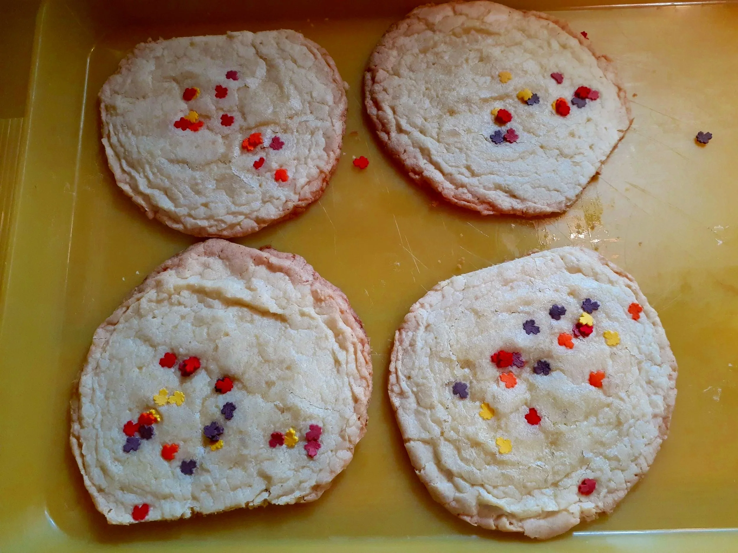 Four round butter sugar cookies with colorful sprinkles on top, placed on a yellow baking tray.