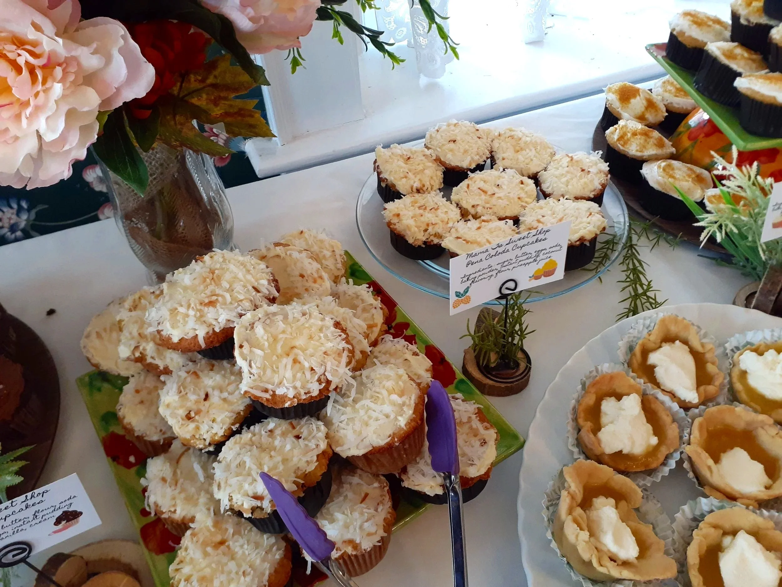 An assortment of cupcakes with white frosting and shredded coconut on top, displayed on colorful plates at a dessert table. A flower arrangement is visible in the background.