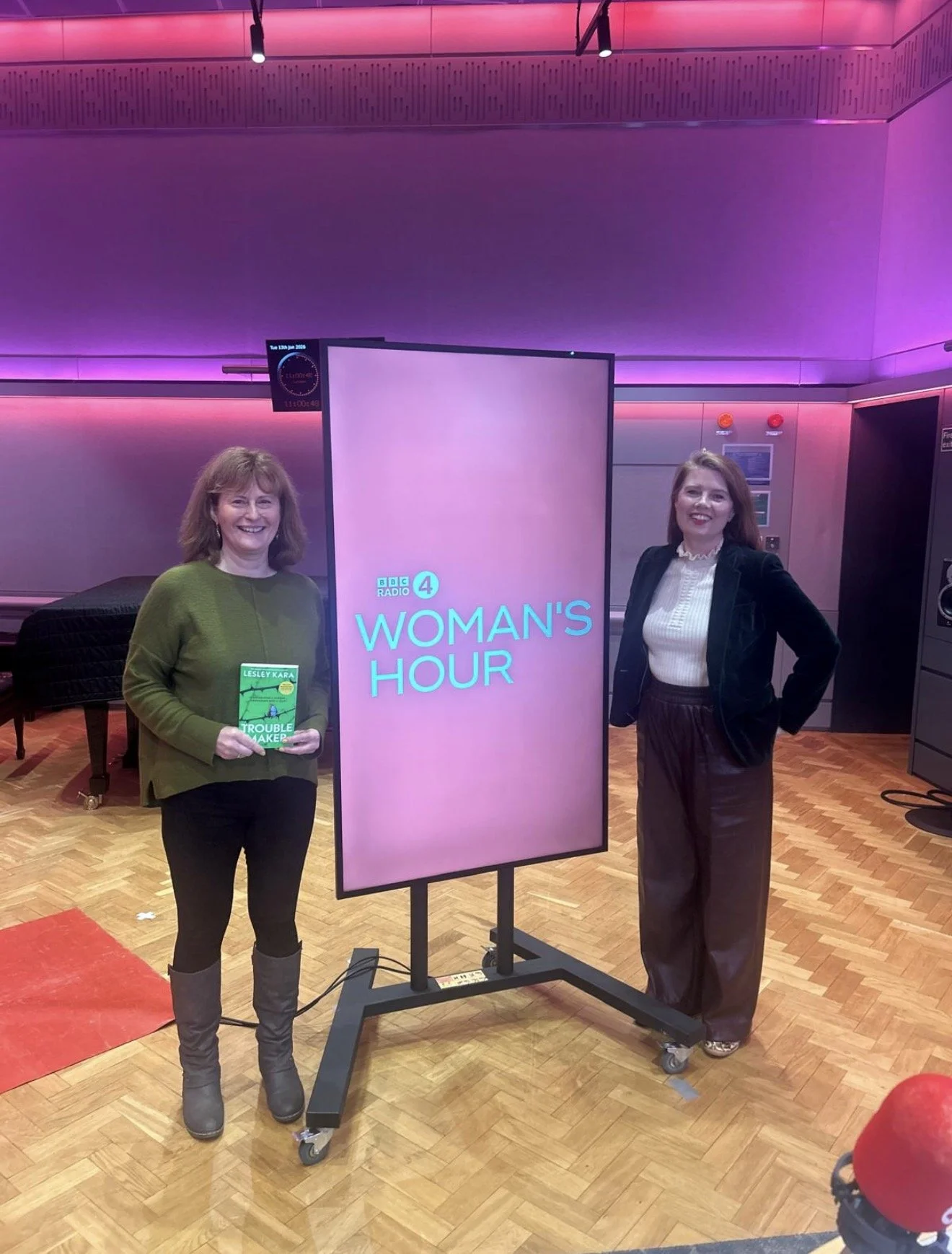 This is a picture of the broadcaster Nuala McGovern and the author Lesley Kara standing next to the BBC Radio 4 Woman's Hour banner. Lesley is holding a copy of her book, Troublemaker.