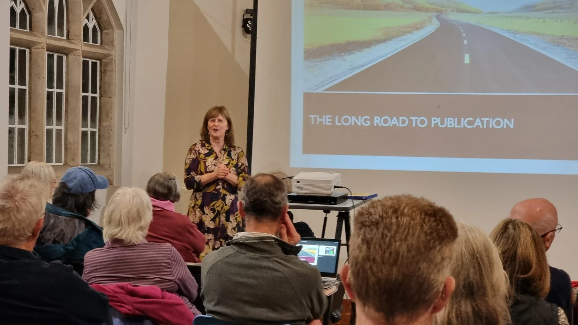 Picture of a woman standing in front of an audience and next to a powerpoint presentation entitled The Long Road To Publication