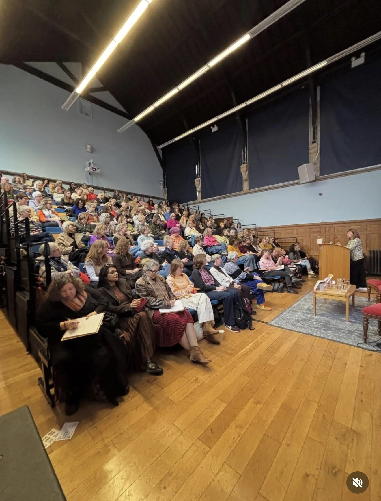 Picture of author Lesley Kara standing at a lectern in front of a packed lecture theatre/auditorium and delivering a talk