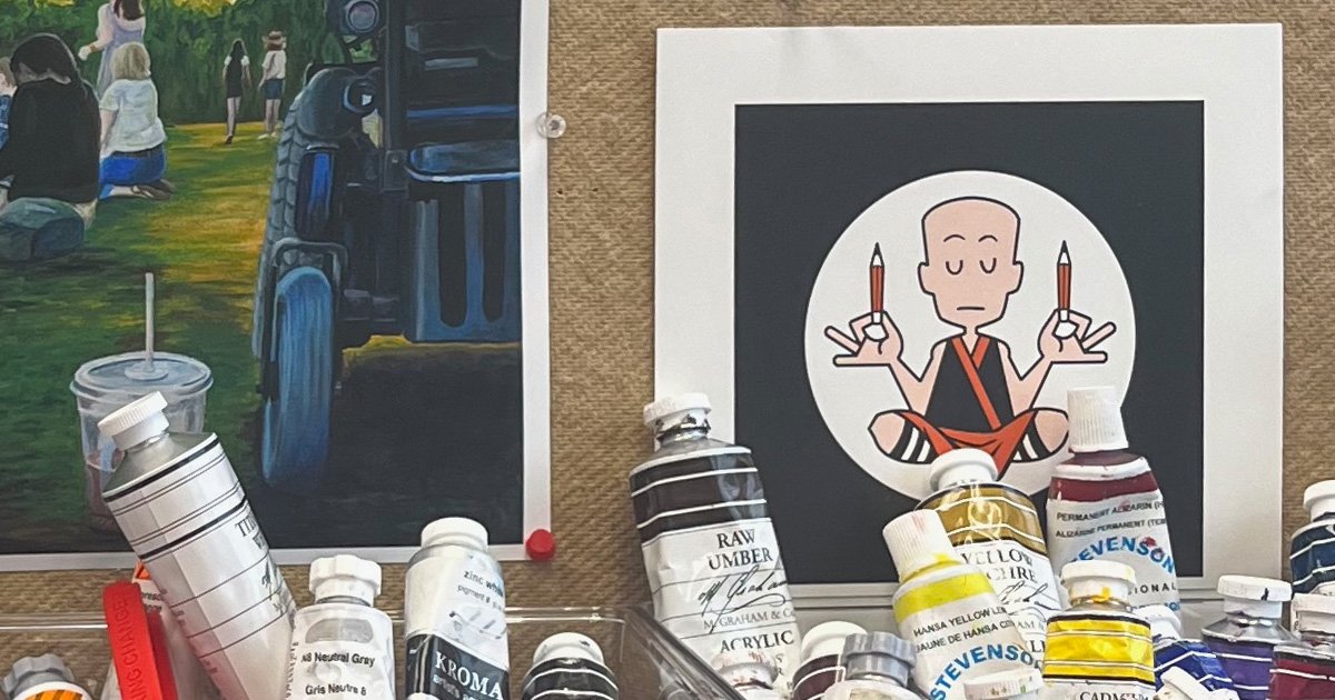 Bulletin board sitting behind a collection of paint tubes. On the bulletin board is pinned an illustration of a monk seated cross-legged holding two pencils up vertically. The print is from the Zen Pencils webcomic.
