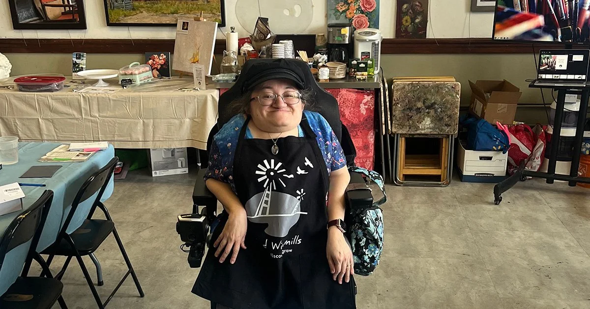 Athena seated in her wheelchair in an art studio. She's smiling and wears a black painter's apron over a flowered blouse.