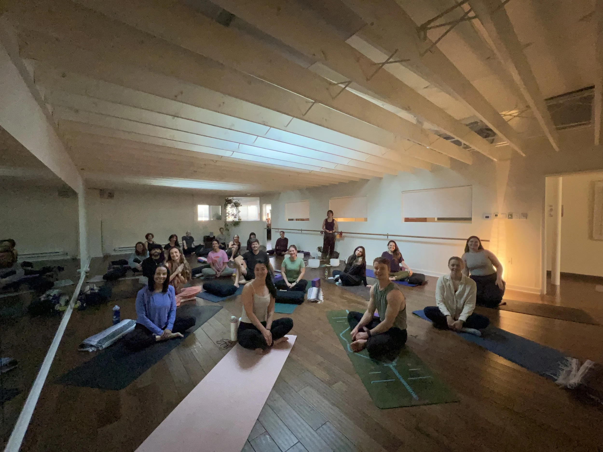 A group of people sitting cross-legged on yoga mats in a spacious, softly lit studio with a high wooden ceiling, some holding meditation cushions, and a woman standing at the front leading a class.