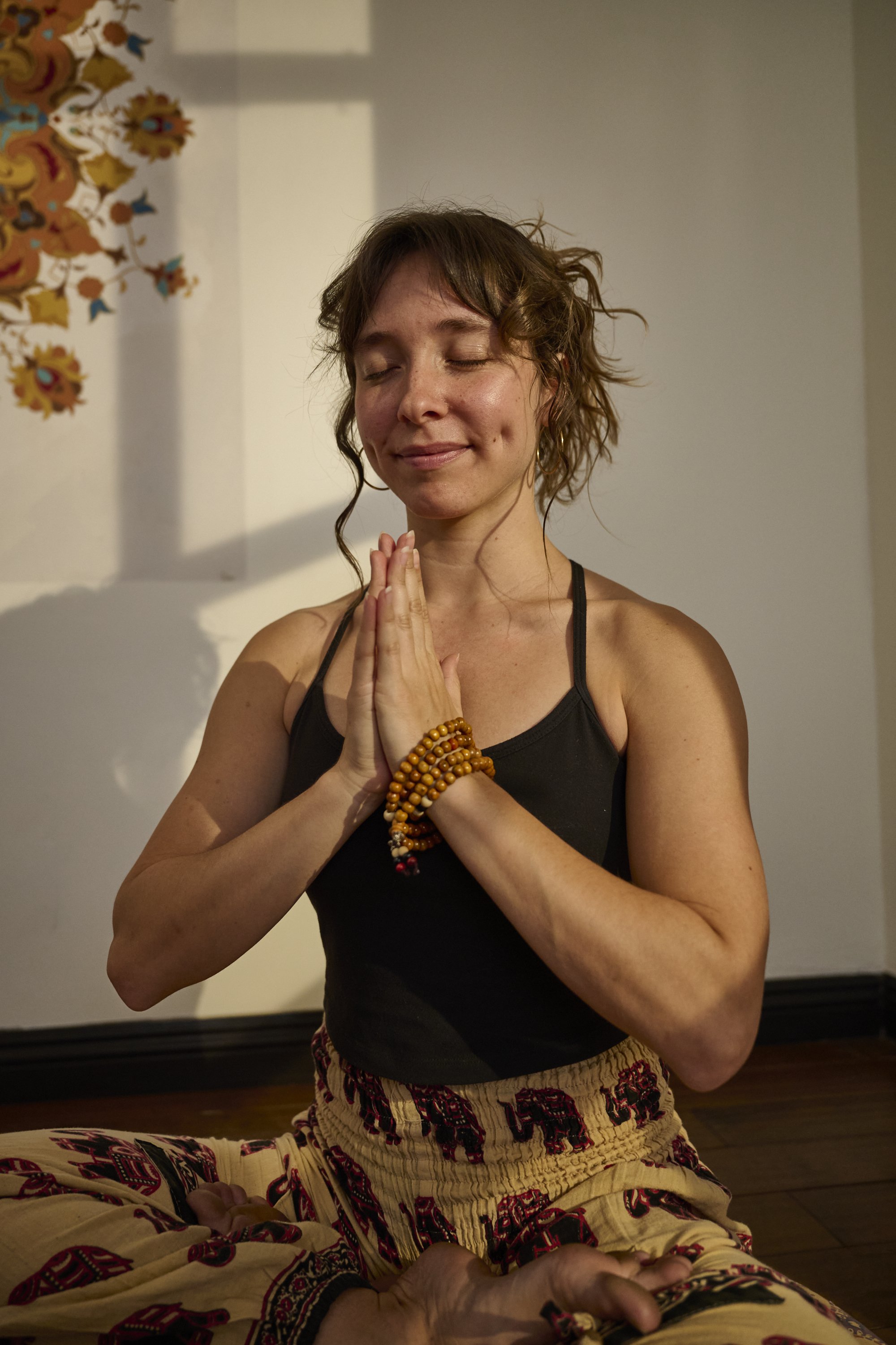 A woman with closed eyes and a gentle smile, in a prayer pose with hands together, wearing beaded bracelets, black tank top, and patterned pants, sitting indoors with a colorful tapestry on the wall.