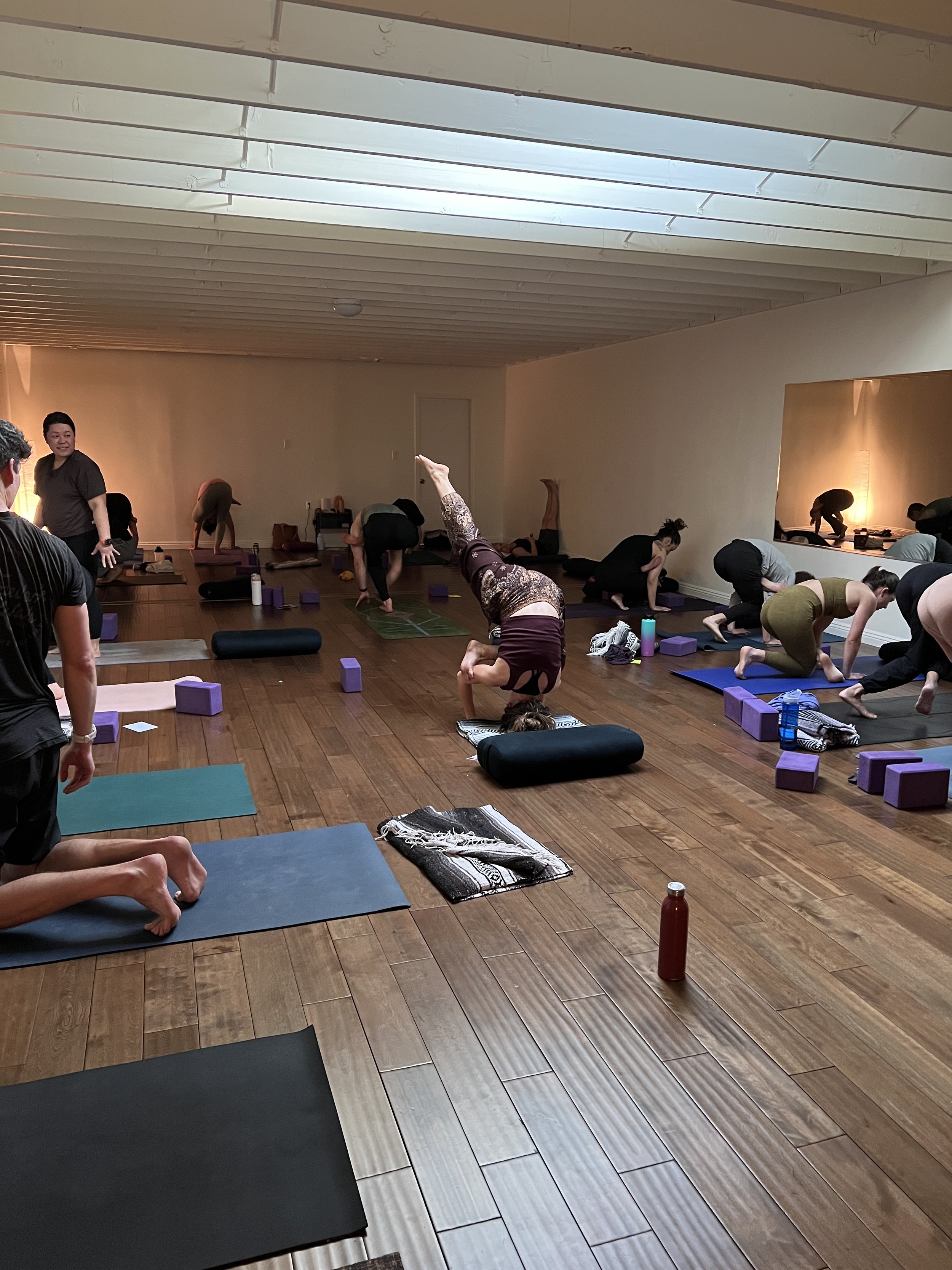 A group of people participating in a yoga class in a room with wooden floors and minimalist decor. Participants are on yoga mats, some in poses, with yoga blocks and water bottles nearby.