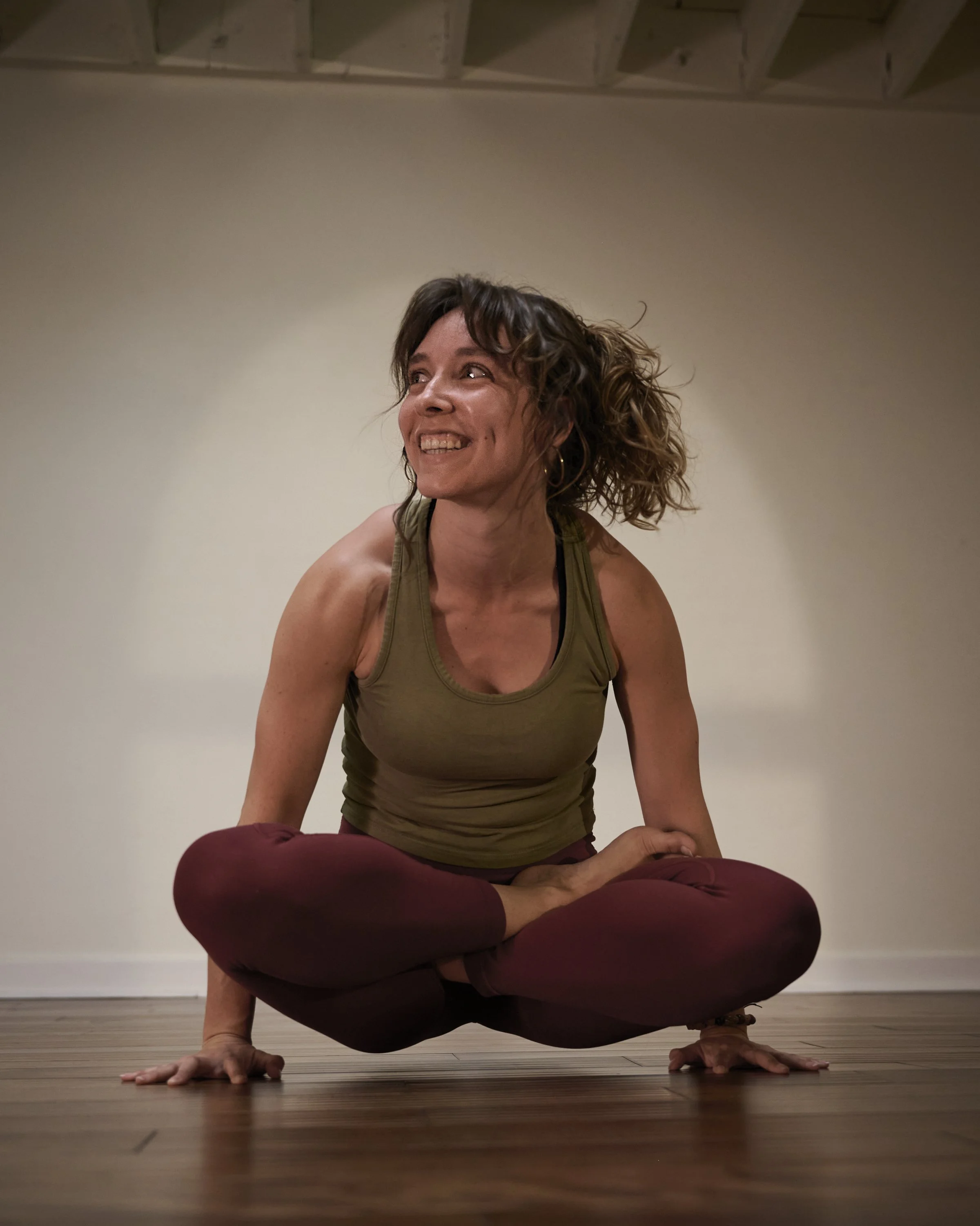 A woman practicing yoga, balancing on hands with knees raised and crossed, on a wooden floor against a plain wall.
