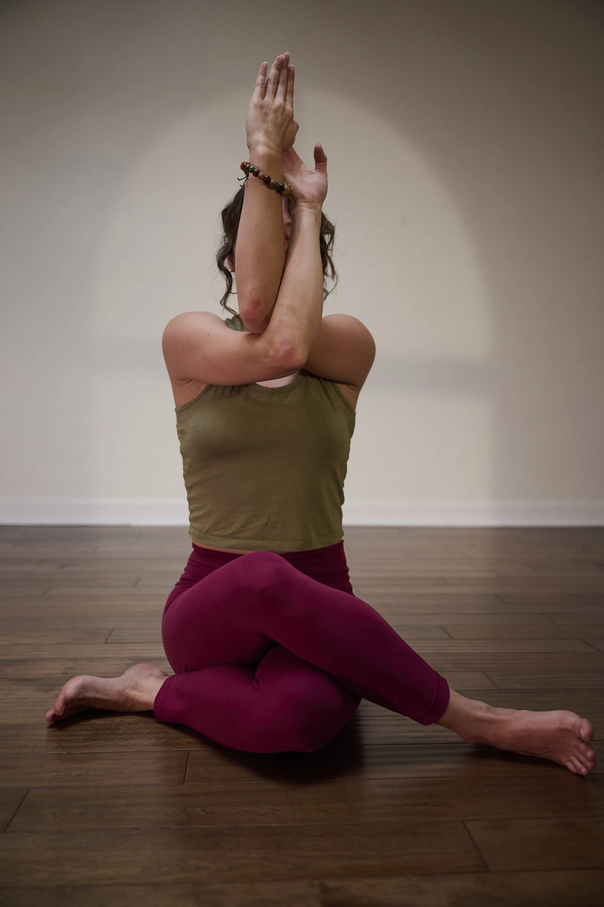 A woman performing a yoga pose on a wooden floor, sitting on her legs with one leg bent and the other extended outward, and her hands in a prayer position above her head. She is wearing a tan sleeveless top and maroon leggings, with a beaded bracelet on her wrist.