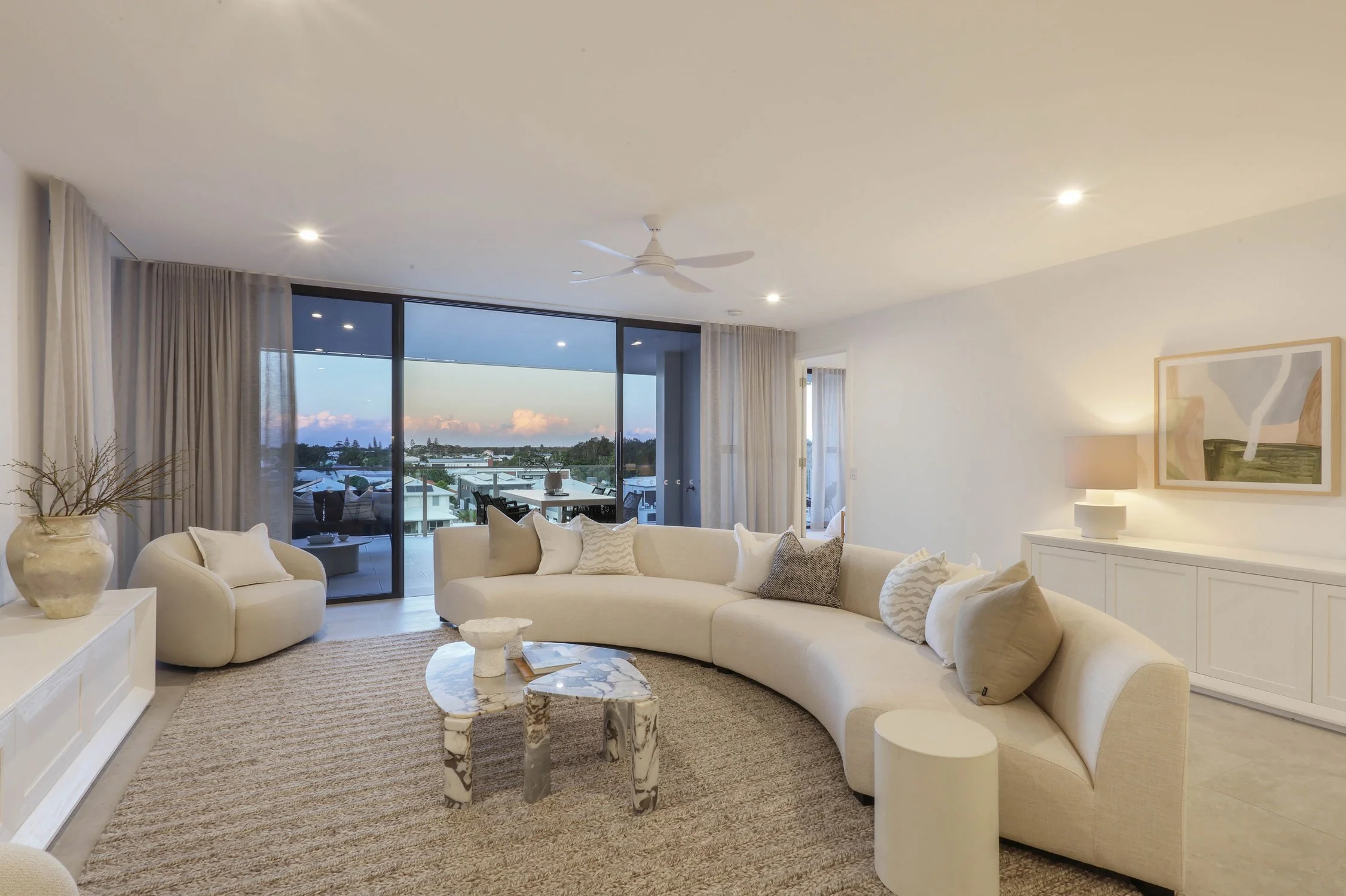 Bright living room with curved white sofa, marble coffee table, beige armchair, and sliding glass doors leading to balcony with outdoor seating and a view of the sky and neighborhood. luxury penthouses for sale In Australia