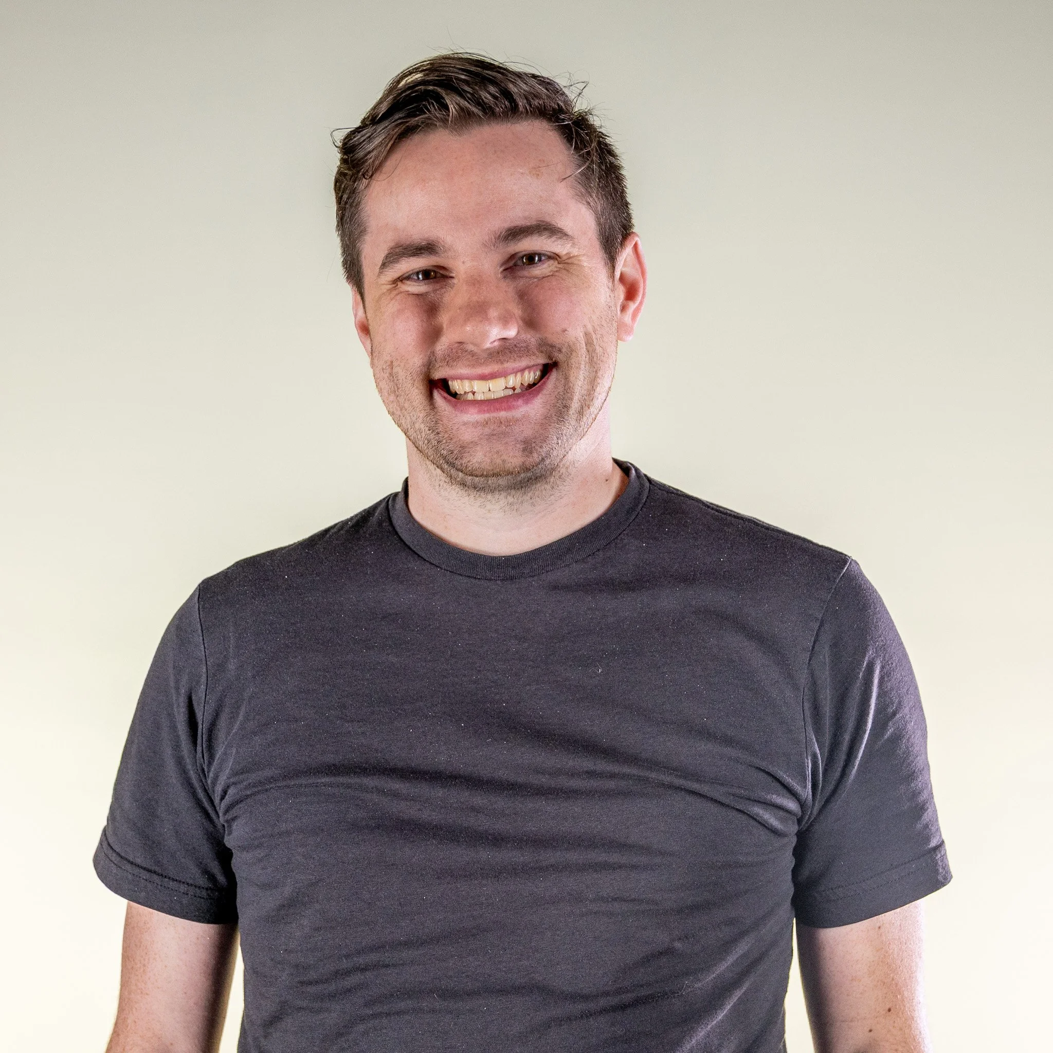 Close-up of a smiling young man with short dark hair, wearing a dark grey T-shirt, against a plain light background.