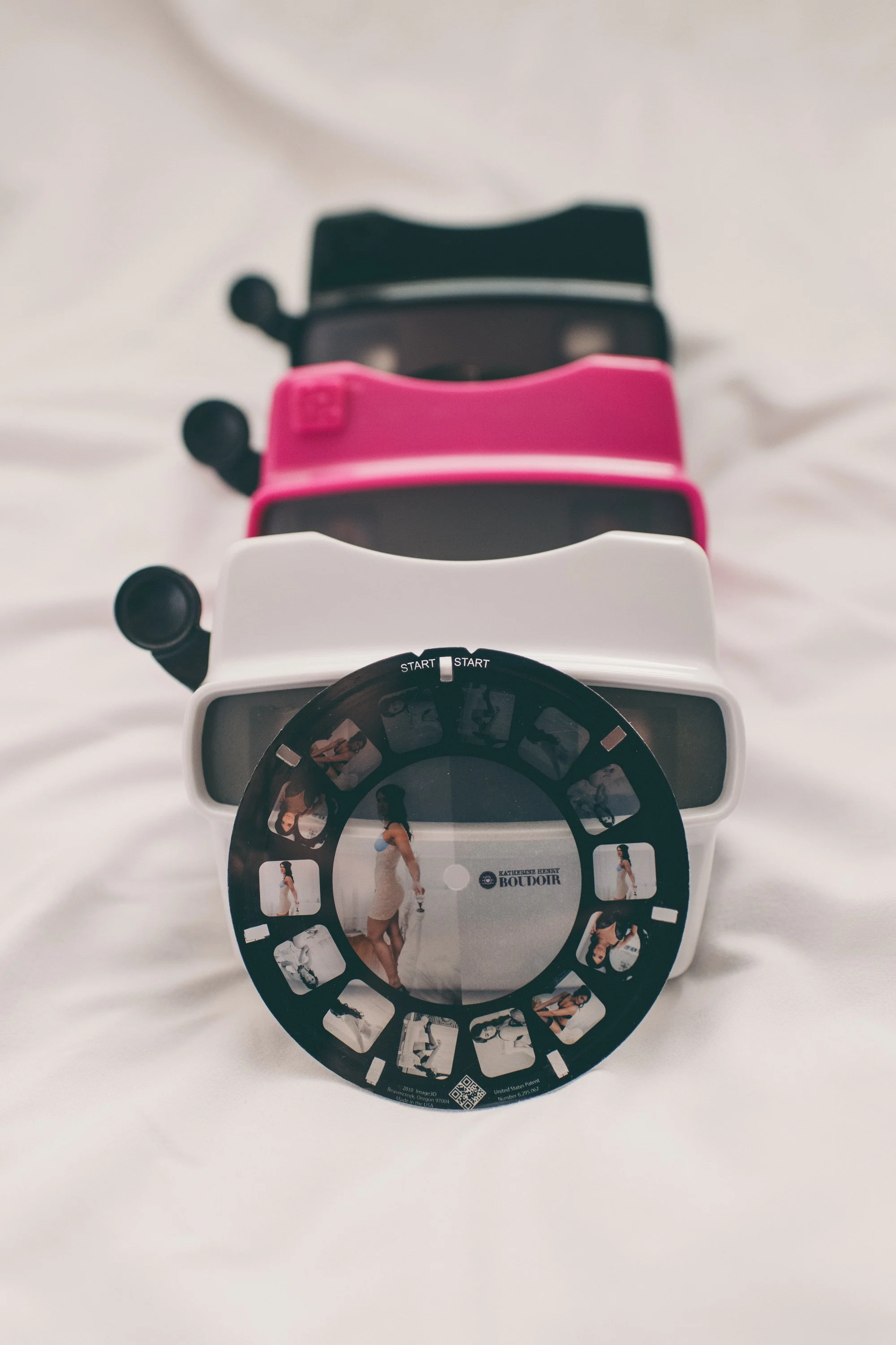 A collection of colorful viewmasters, including black, pink, and white, with a round slide featuring images of women in various poses placed in front of the containers.