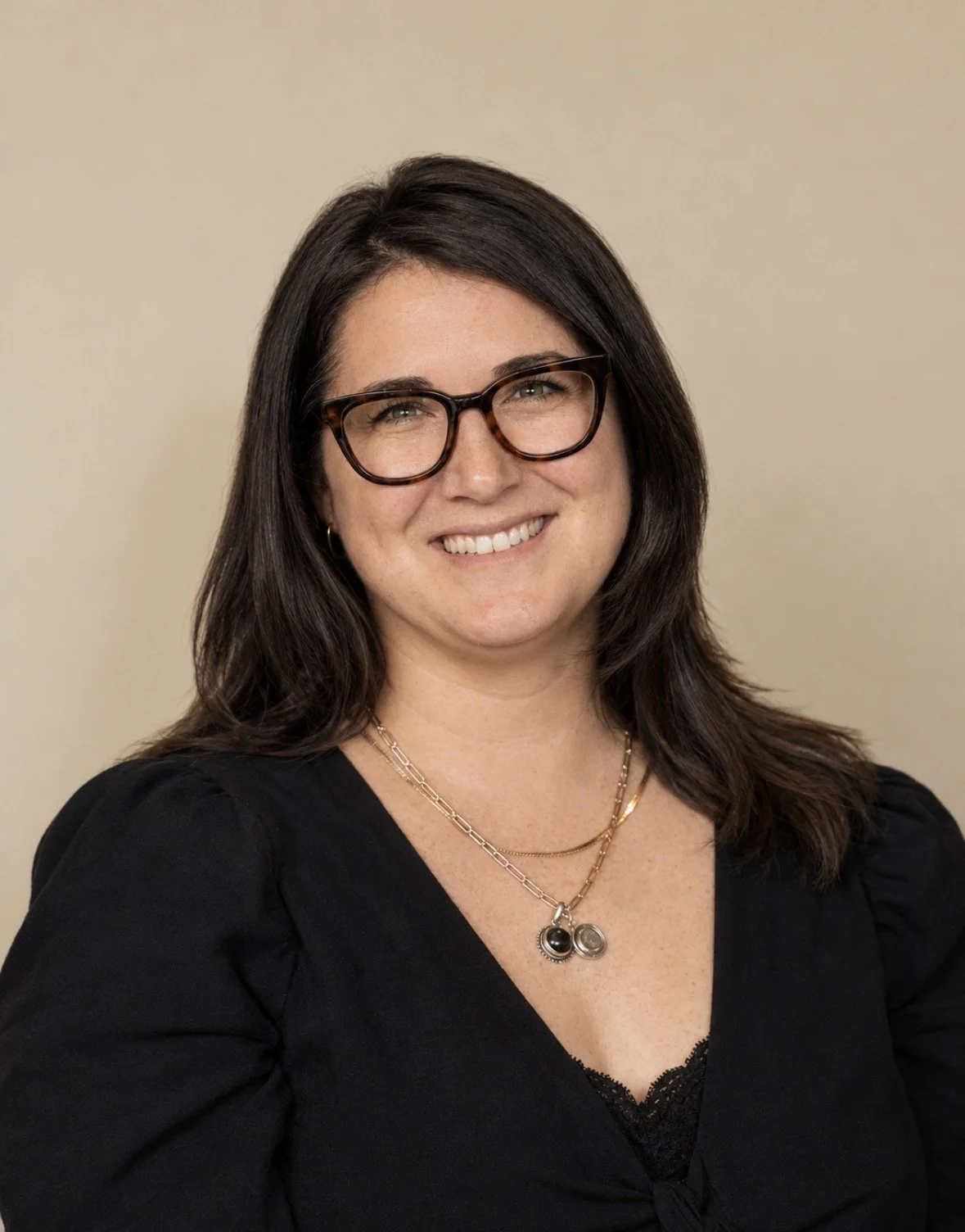 Professional headshot of a smiling woman with dark hair and glasses wearing a black top and layered necklaces