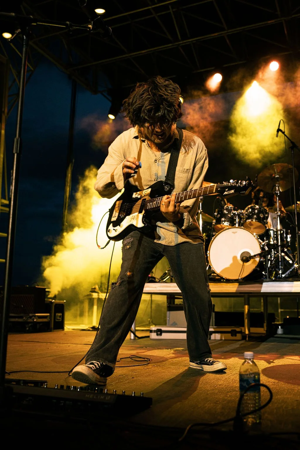 A musician playing electric guitar on stage during a live concert at night, with drummer in the background and colorful stage lights.